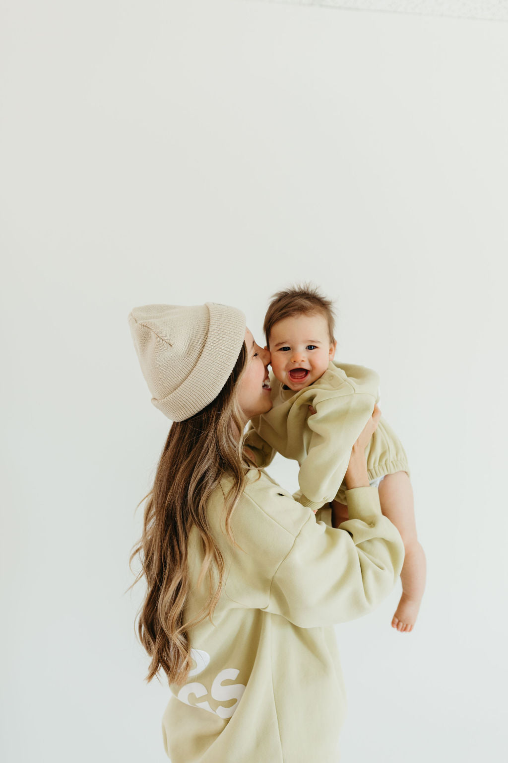 A woman wearing a beige beanie holds a smiling baby dressed in the "All The Good Things" sweatshirt romper from forever french baby. She gently kisses the baby's cheek against a plain, light-colored background.