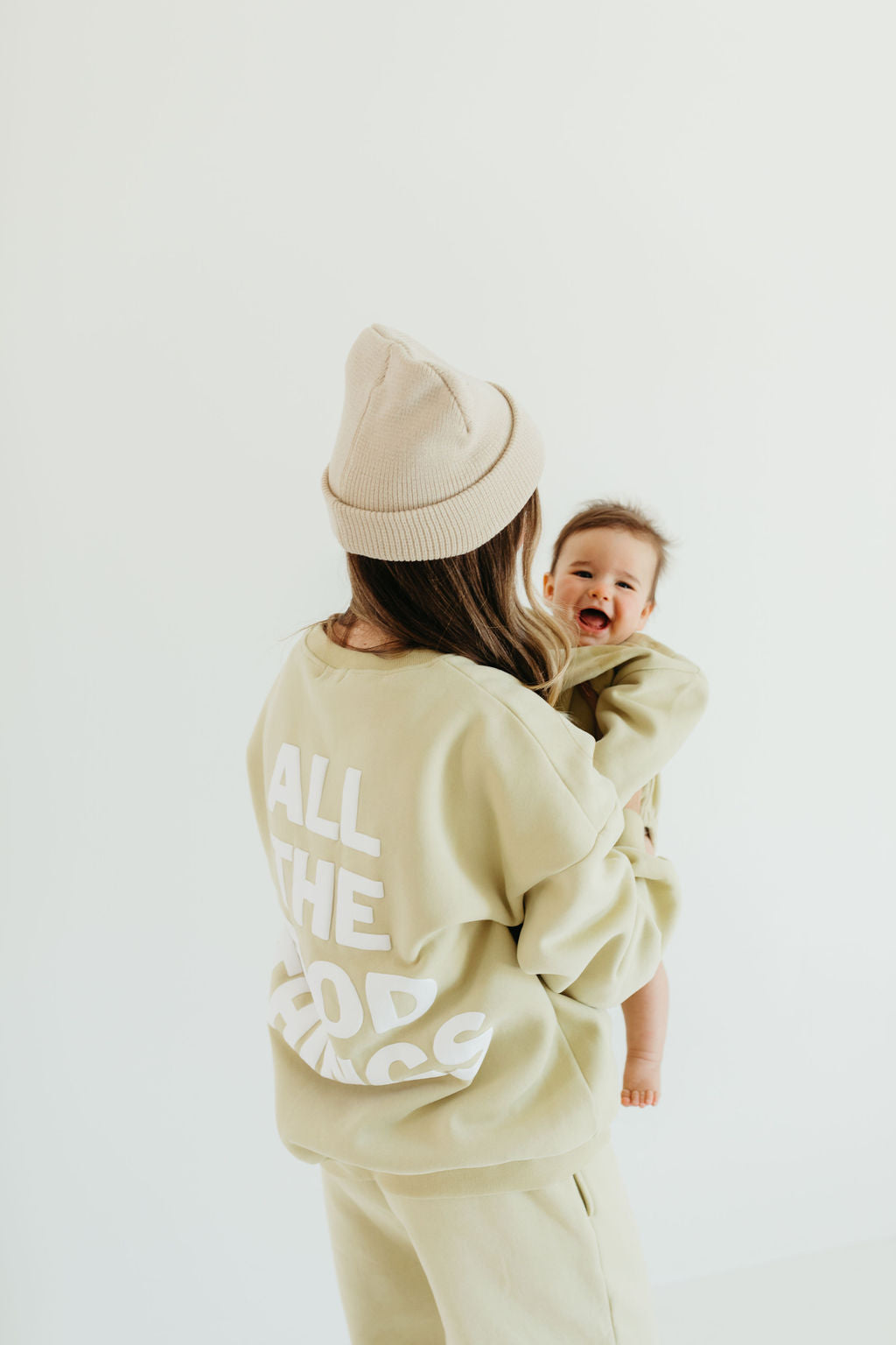 A woman in an avocado green beanie and a sweatshirt with the phrase "All the Good Things" is holding a smiling baby. Both are dressed in matching beige sweat sets, capturing the cozy charm of Forever French Baby against a plain white backdrop.