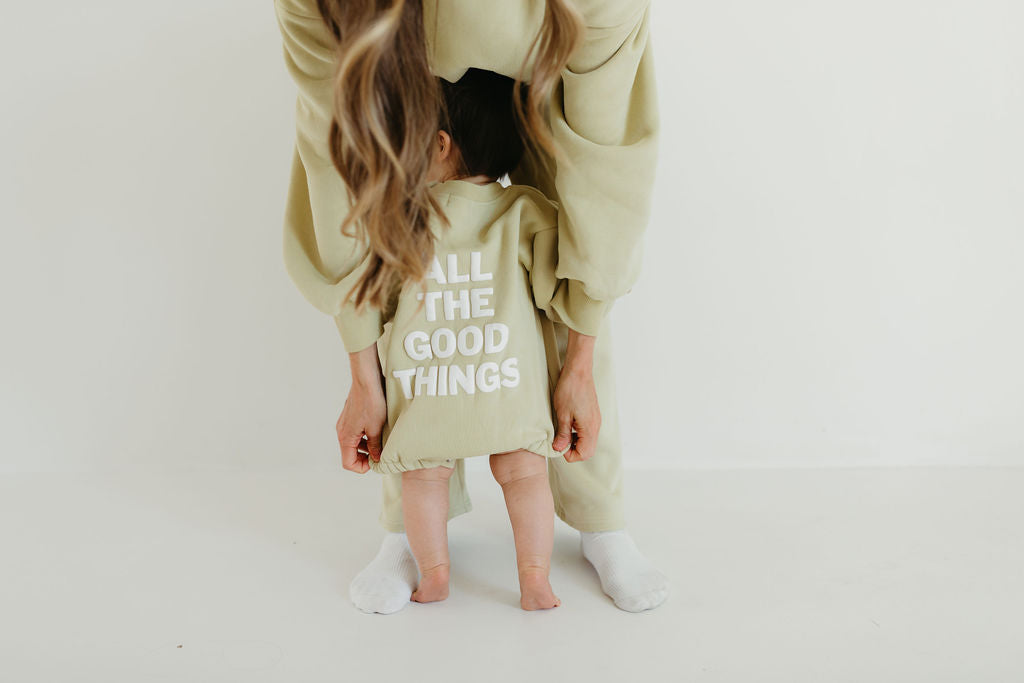 A parent dressed in an avocado green outfit bends over to adjust their toddler's matching "All The Good Things" sweatshirt romper by forever french baby, capturing a sweet moment in a minimal, light-colored room.