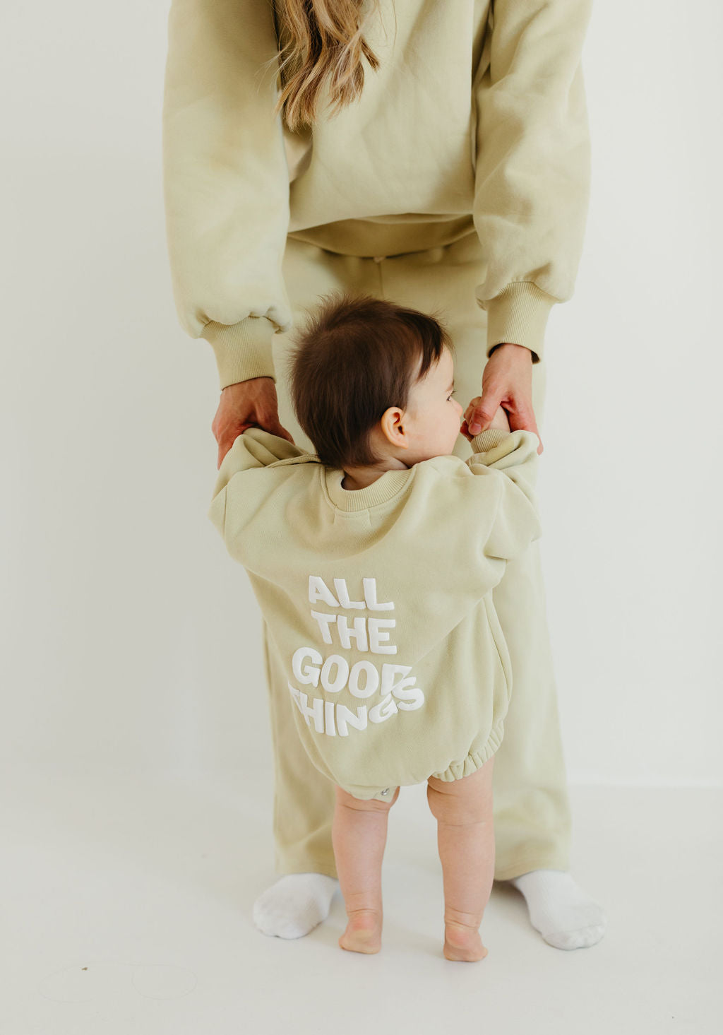 An adult holds a child's hands as the child stands, both dressed in matching beige sweat sets from the Forever French Baby brand. The child's sweater from their collection features "All the Good Things" written on the back in white text.