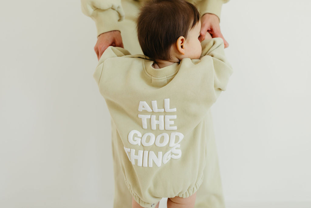 A baby, clad in the oversized Sweatshirt Romper "All The Good Things" by forever french baby, faces away from the camera. This adorable moment captures the essence of Forever French Baby style as the little one clings to an adult's hands in a softly lit room.