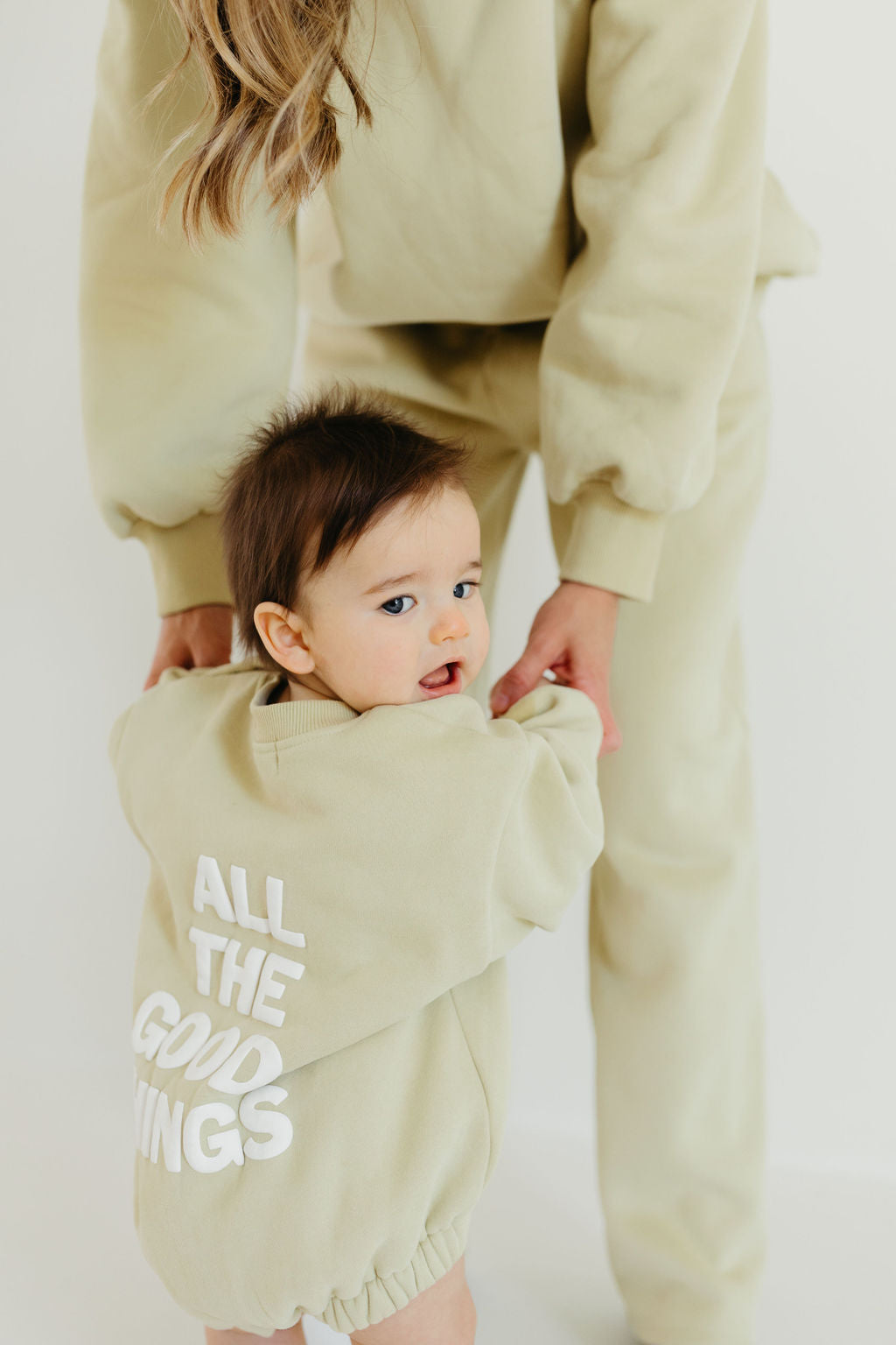 An adult, wearing the "Adult Sweat Set | All the Good Things" by Forever French Baby, helps a toddler who is sporting an avocado green sweatshirt with "ALL THE GOOD THINGS." Both are adorned in matching cozy sweat sets. The minimalist scene exudes the charm of Forever French Baby's design.