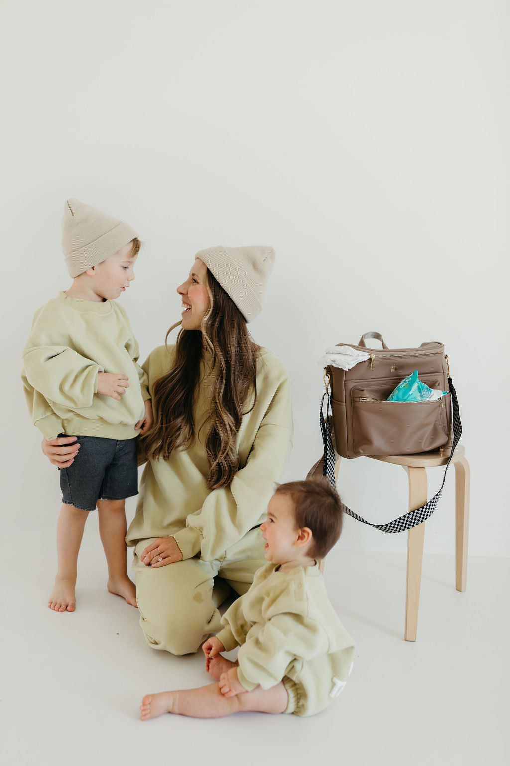 A woman and two young children, all wearing beige outfits, pose indoors. The woman sits on the floor alongside a black checkerboard rug, smiling at the standing child next to her while the seated child looks up at them. A stylish taupe ff Diaper Bag by forever french baby, featuring insulated bottle pockets, sits on a nearby stool.