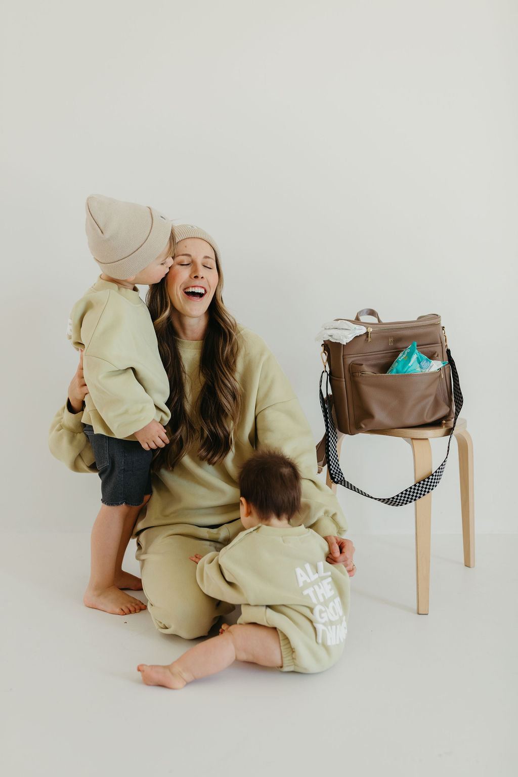 A woman, joyfully laughing, sits with two children dressed in matching beige outfits. One child stands close to her, hugging tightly, while the other sits on the floor. Next to them on a chair is a Taupe "ff Diaper Bag" from forever french baby, adorned with vegan leather accents and printed text, filled with items inside.