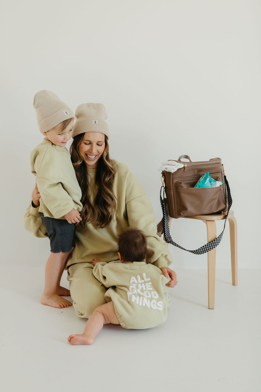 A smiling woman kneels beside two young children, all dressed in matching beige outfits. One child stands while the other sits on her lap. Nearby, a taupe ff Diaper Bag from forever french baby rests on a stool, partially filled with items and featuring insulated bottle pockets. The child's back features the text "ALL THE GOOD THINGS.