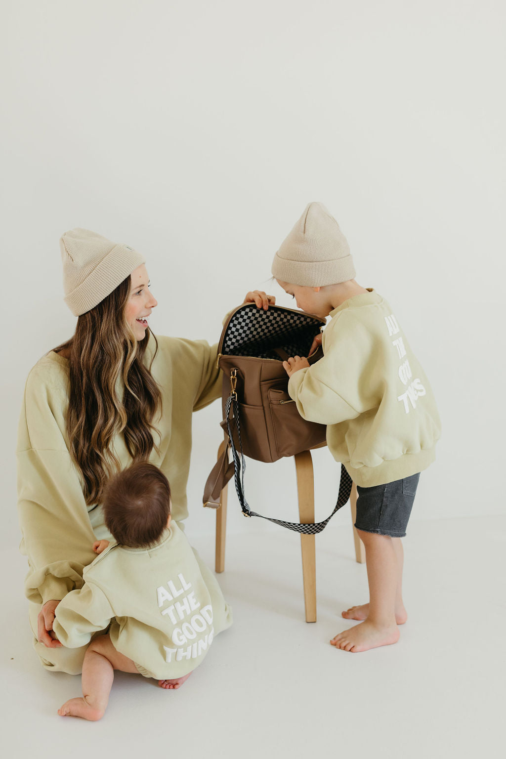 A woman and two children, all wearing matching beige hats and light green sweatshirts with text on the back, sit together. One child stands on a stool peering into a forever french baby ff Diaper Bag in Taupe, which features a black checkerboard design. The setting is bright and minimalist.