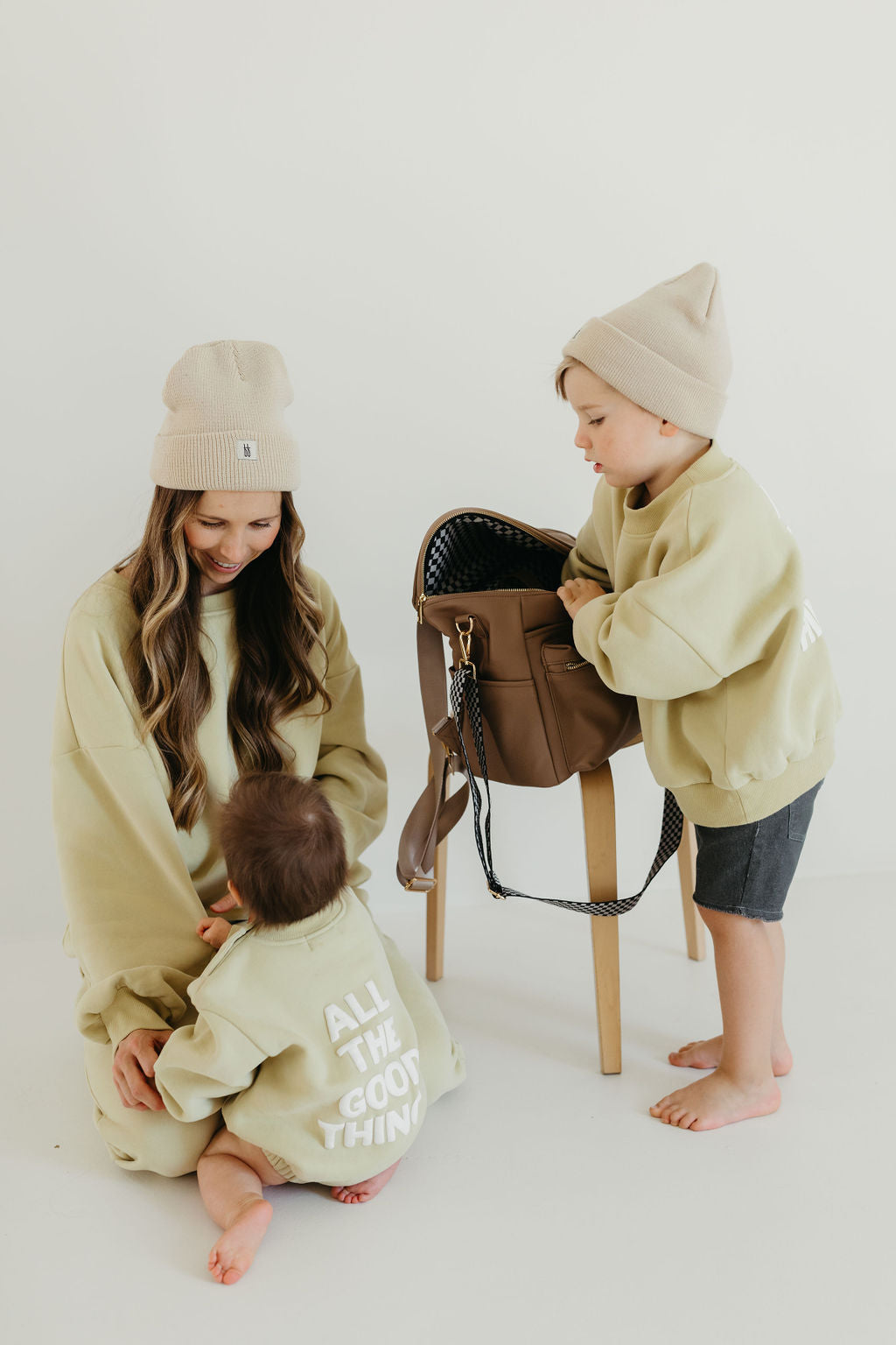 A woman and two children in matching beige outfits and beanies are interacting. One child, holding a forever french baby ff Diaper Bag | Taupe, sits on a stool, while the other is on the floor. The woman's sweatshirt reads, "All the good things.