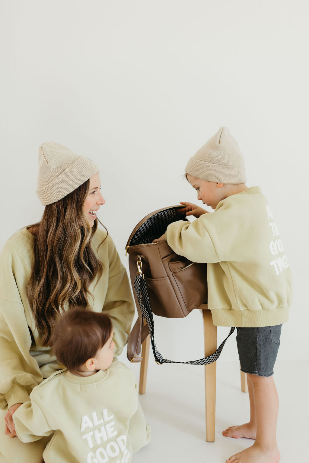 A woman and two children, all dressed in matching light beige sweaters with "ALL THE GOOD" text on their backs and wearing knit hats, are gathered around a chair. One child is peering inside a taupe ff Diaper Bag by forever french baby placed on the chair.