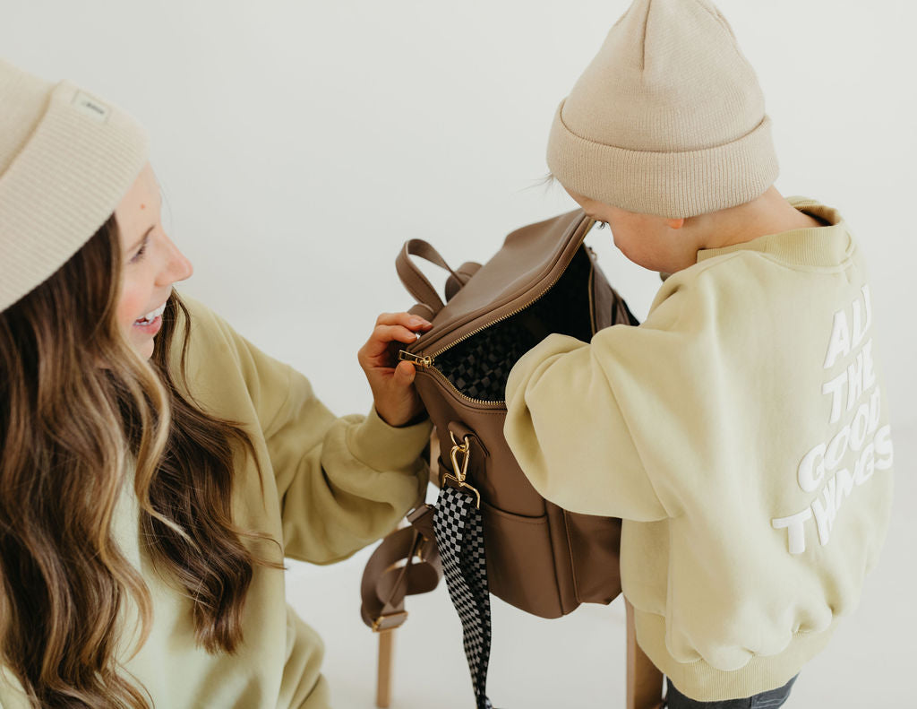 A woman and a child, both wearing beige beanies and green sweatshirts with black checkerboard patterns that read "ALL THE GOOD VIBES," are sitting together cheerfully. The child is happily opening the ff Diaper Bag in Taupe by forever french baby, which features insulated bottle pockets.