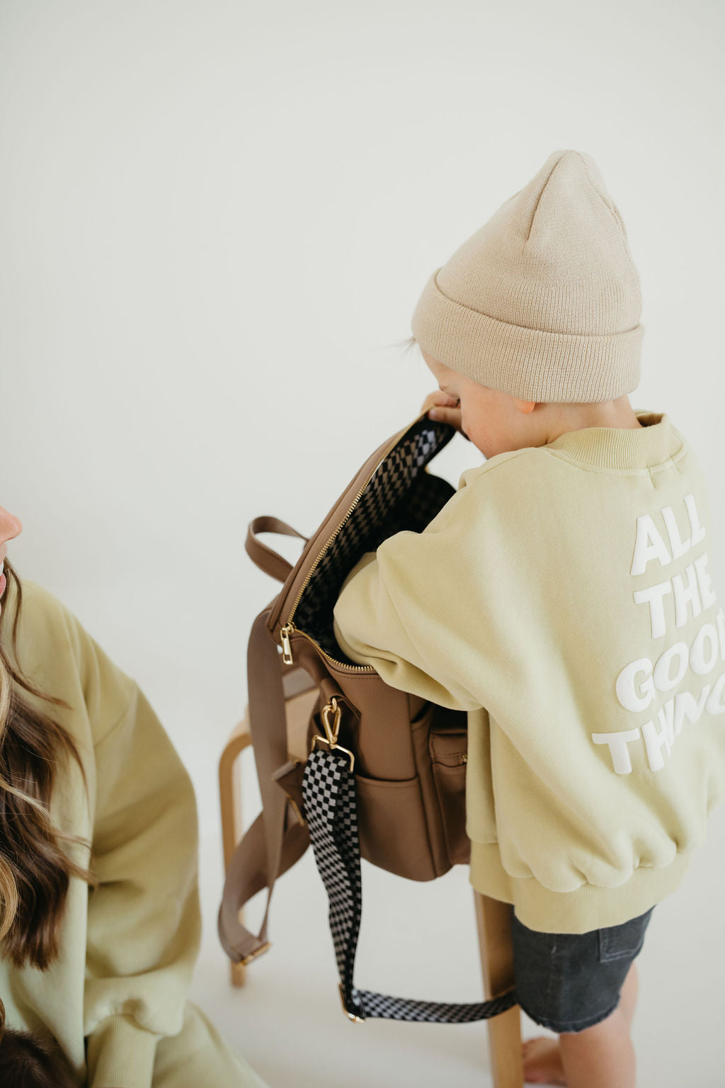 A child wearing a beige beanie and an oversized sweatshirt with "ALL THE GOOD THINGS" on the back stands on a chair, reaching into a forever french baby's ff Diaper Bag in taupe with vegan leather accents. Another person is partially visible, sitting nearby in the minimal and neutral setting.