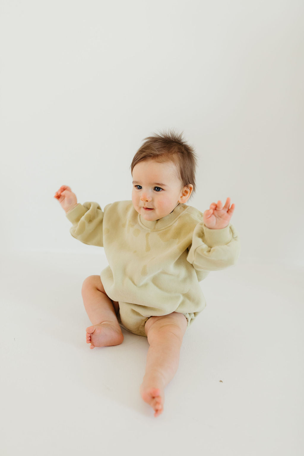 A baby in an avocado green "All The Good Things" sweatshirt romper from Forever French Baby sits on a plain white background, raising both arms with a curious expression. The baby's hair is slightly tousled, and the soft light creates a gentle atmosphere perfect for the Forever French Baby collection.