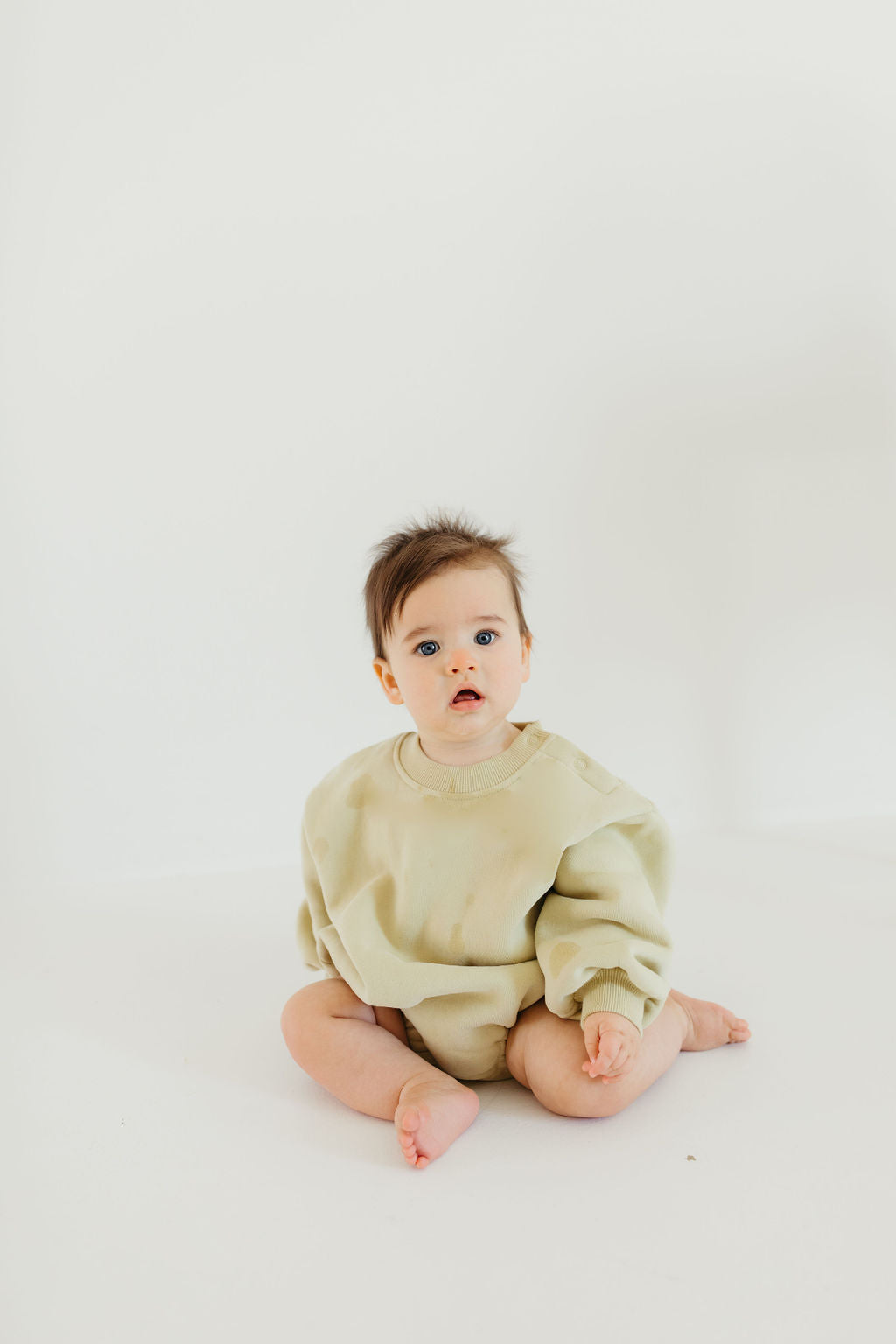 A baby in the 'All The Good Things' sweatshirt romper by forever french baby sits on a white surface against a plain background, looking slightly to the side. The child has dark hair, a curious expression, and exudes the timeless charm associated with forever french baby.