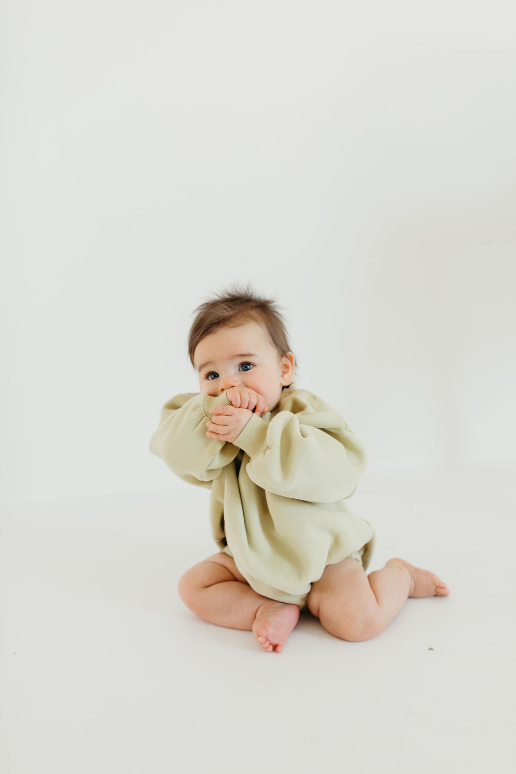 A baby with light brown hair is sitting on the floor, dressed in a fashionable avocado green "All The Good Things" sweatshirt romper from Forever French Baby. Their hands are raised to their mouth against a soft, light cream background.