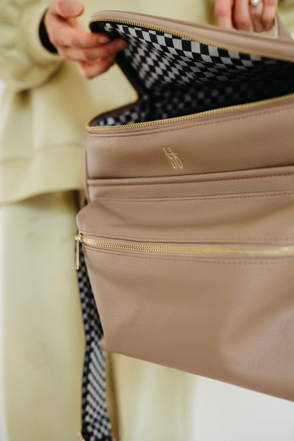 Dressed in a beige outfit, a person opens the taupe ff Diaper Bag by forever french baby, featuring gold zippers and revealing a black checkerboard interior, highlighting the bag's stylish design and functionality.