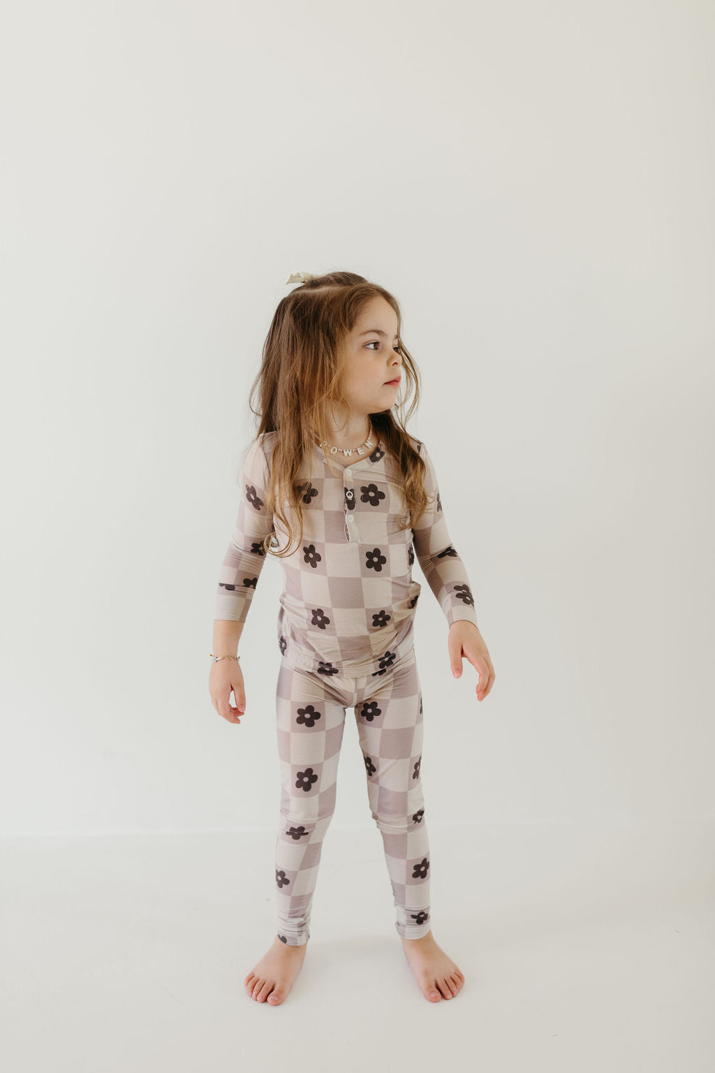 A young girl with long hair stands barefoot against a white wall, wearing the Bamboo Two Piece Pajamas in the Flower Child design by forever french baby. The pajamas feature checkered patterns with floral designs and are made from breathable fabric. She looks to the side with a curious expression.