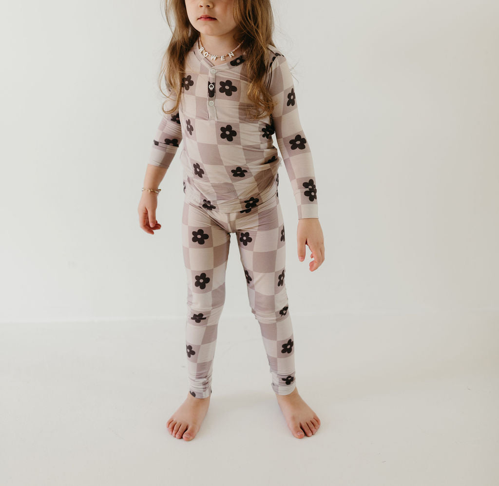 A child is seen barefoot on a white floor, dressed in the "Bamboo Two Piece Pajamas | Flower Child" by forever french baby. This matching ensemble features a light and dark gingham pattern with floral designs and is hypo-allergenic. Made from breathable fabric, it is ideal for sensitive skin. The child's long hair cascades as they pose against a plain white background.