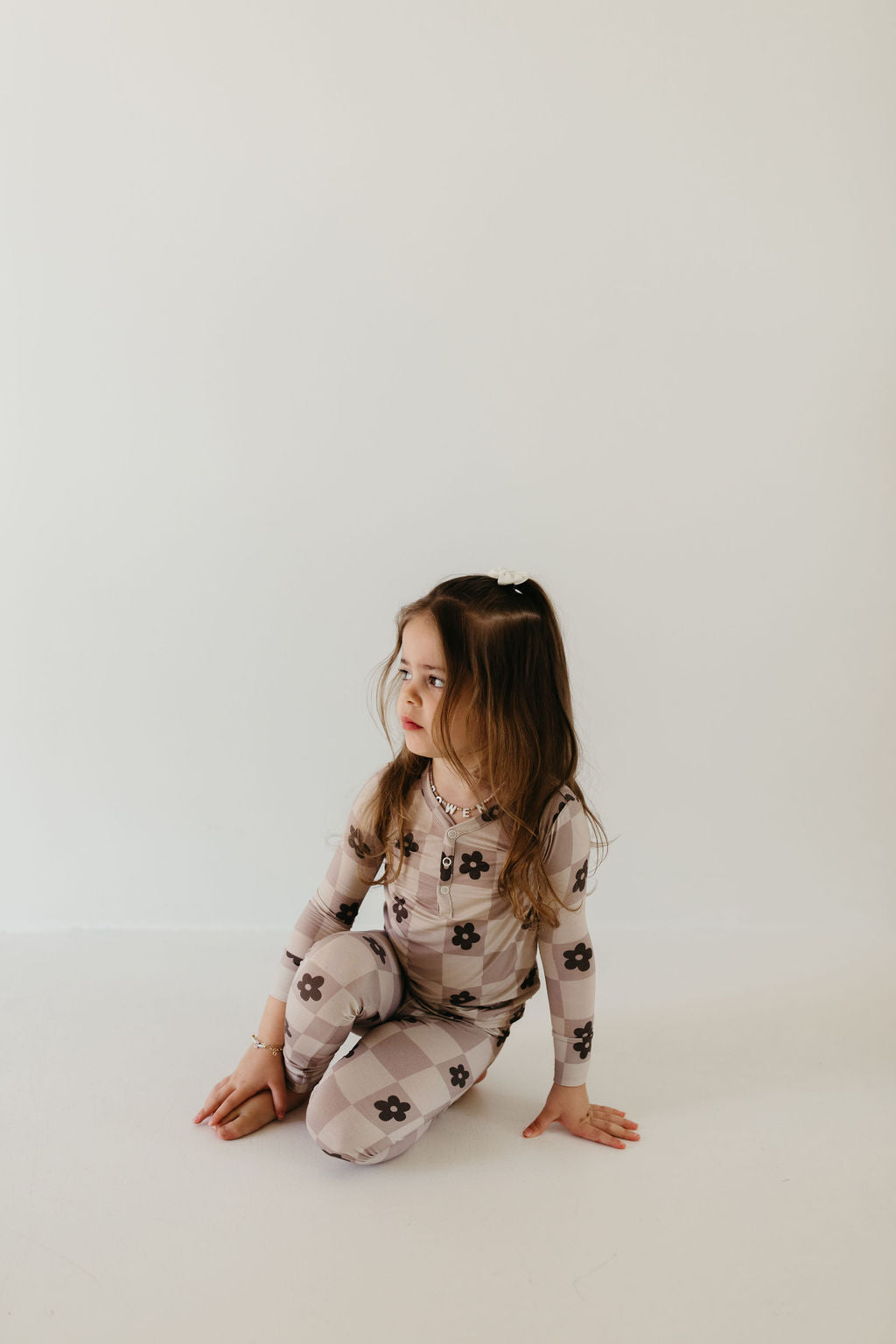 A young girl with long hair sits on the floor against a simple light background, wearing the "Bamboo Two Piece Pajamas | Flower Child" by forever french baby, featuring a beige and brown checkered pattern with floral prints. Her head is turned to the side, and she looks thoughtful in her hypo-allergenic outfit.