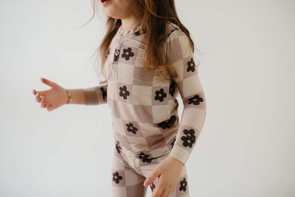 A child models the forever french baby Bamboo Two Piece Pajamas | Flower Child, featuring a beige and brown floral checkered pattern, crafted from breathable bamboo fabric. The child’s long hair flows gently as they stand against a plain white background, with arms slightly outstretched to capture the essence of comfort in hypo-allergenic clothing.