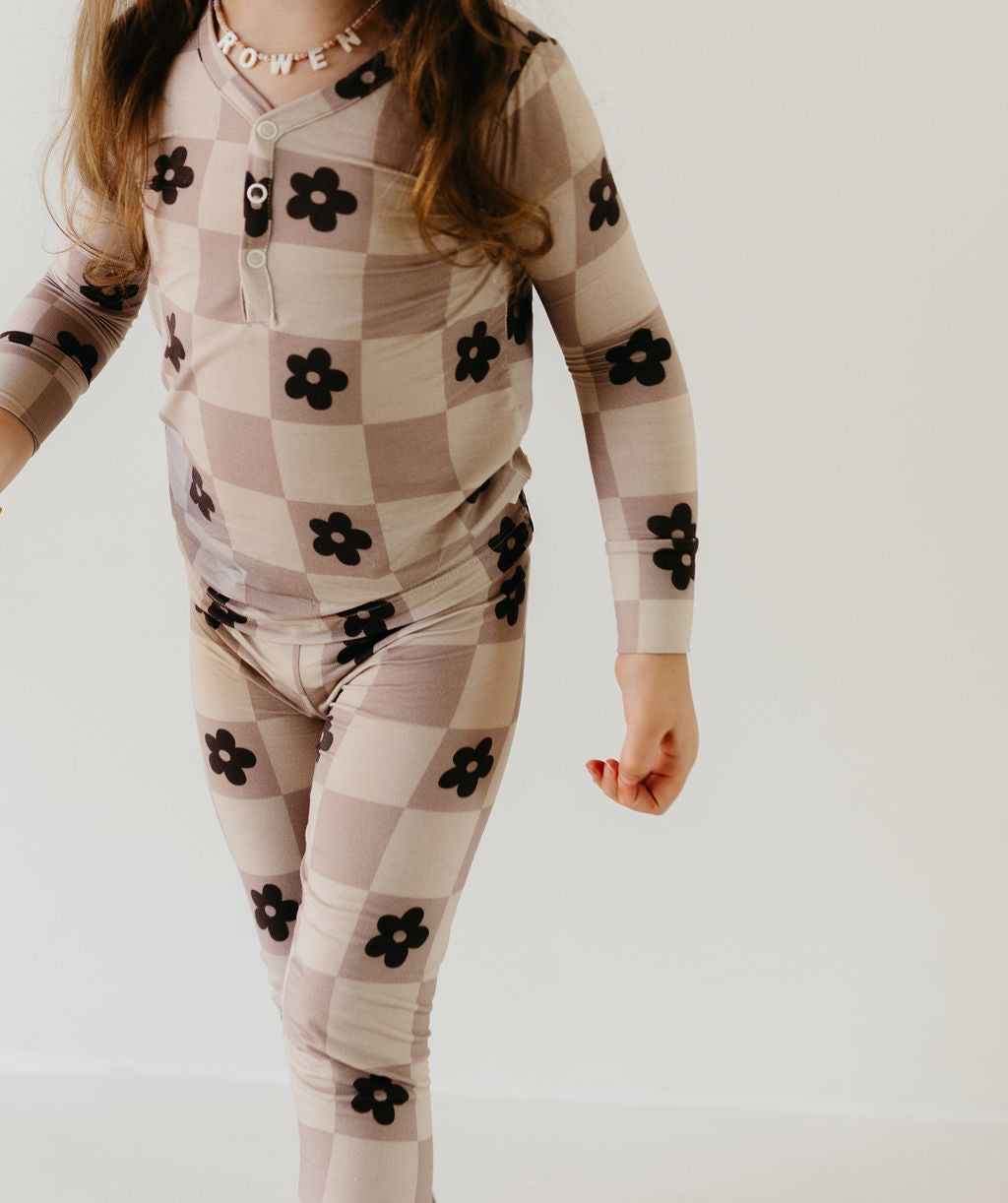 A child is wearing the "Bamboo Two Piece Pajamas | Flower Child" from forever french baby, showcasing a beige and brown checkered pattern adorned with large black flowers. Made from breathable, hypo-allergenic fabric, the set includes a button-up top. The child is posed against a neutral background, embodying both comfort and style.