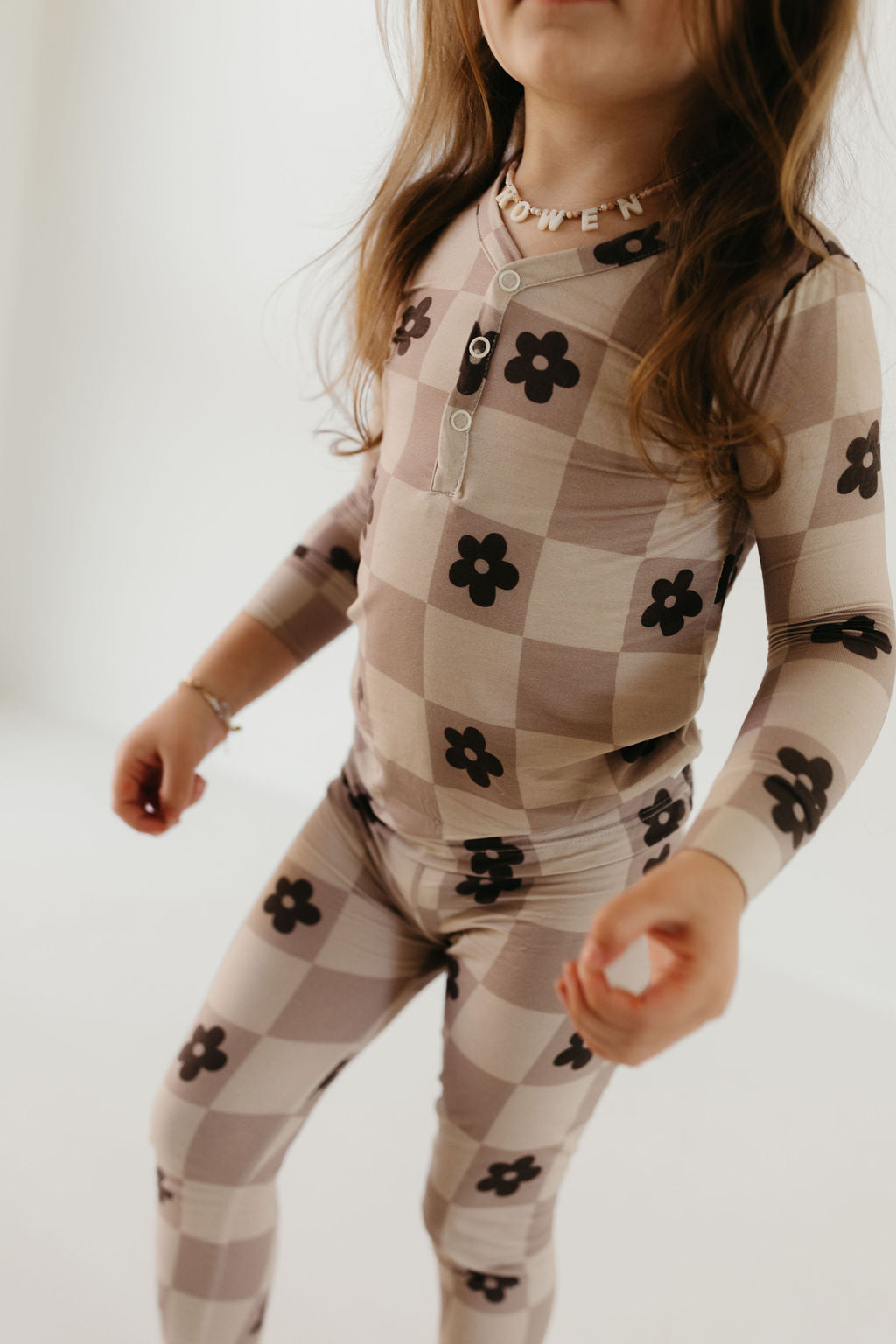 A child is dressed in the Bamboo Two Piece Pajamas from forever french baby, featuring a long-sleeved top and pants crafted from breathable fabric adorned with a beige checkered pattern and dark floral accents. The child's long brown hair complements their personalized beaded necklace, set against a plain light-colored background.