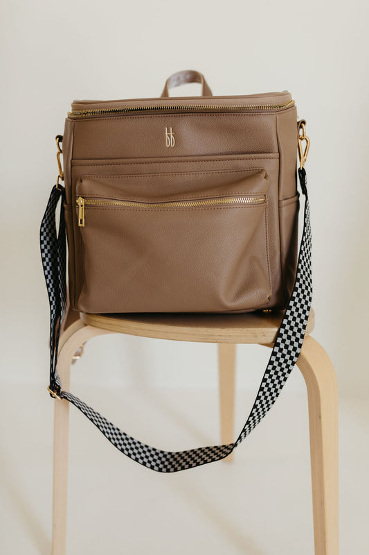 A taupe ff Diaper Bag from forever french baby, featuring gold zippers and a black checkerboard strap, is displayed on a wooden stool. The bag has insulated bottle pockets and boasts a minimalistic design.