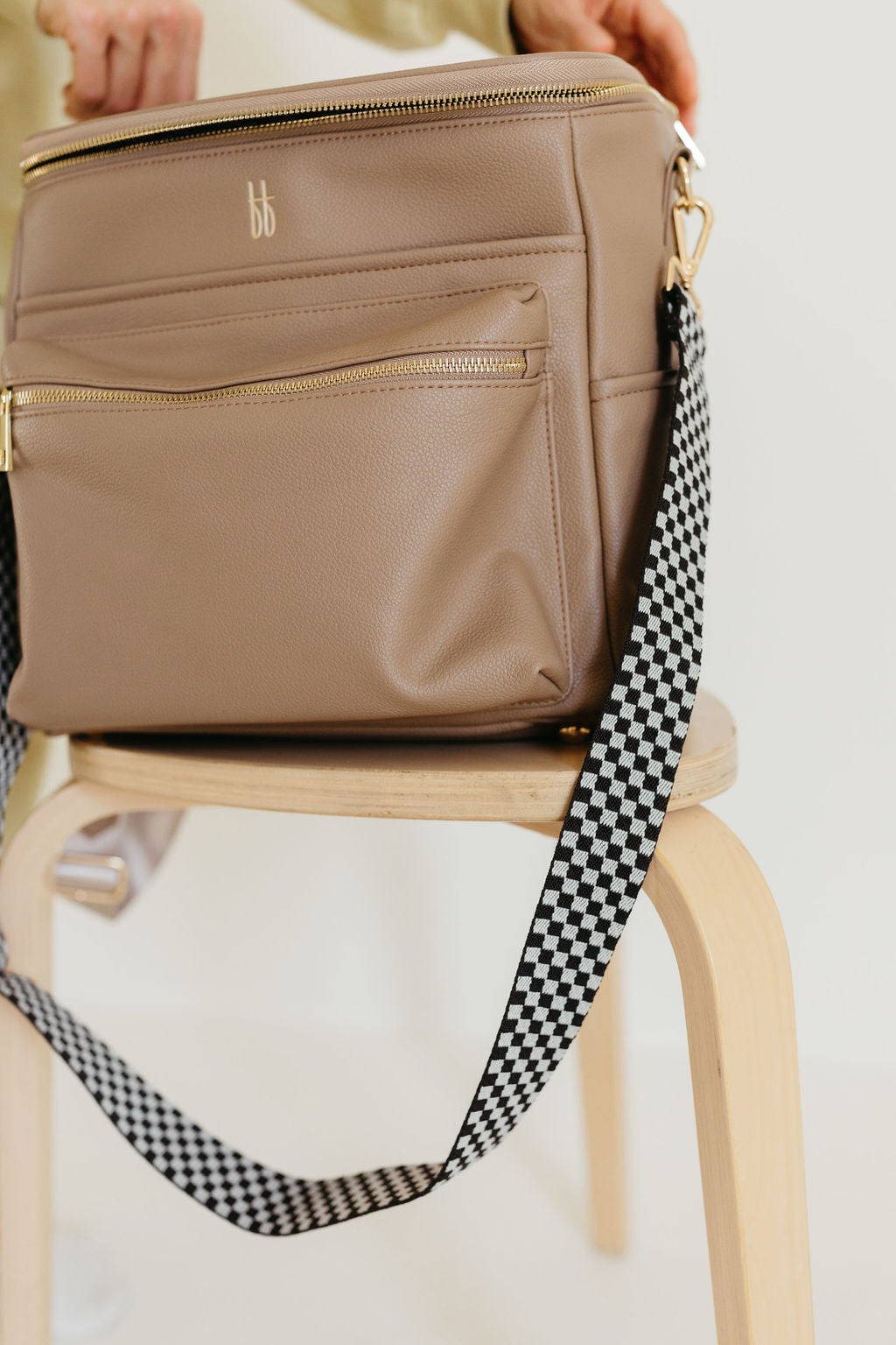 The ff Diaper Bag in Taupe by forever french baby, crafted from vegan leather, features a refined beige color and gold zipper, complete with multiple insulated bottle pockets. It rests on a wooden stool and is adorned with a black and white checkered strap. At the top of the bag, hands are adjusting it against a soft, neutral background.