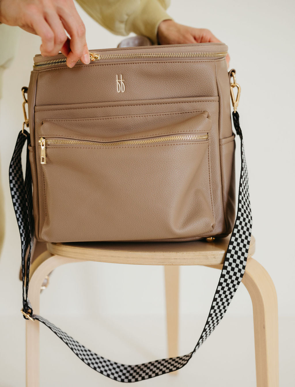 A person opens a taupe ff Diaper Bag by forever french baby, featuring gold zippers, placed on a wooden stool. Made of vegan leather, the bag boasts a black checkerboard lining and is accented by its checkered strap and small "bb" logo on the front.