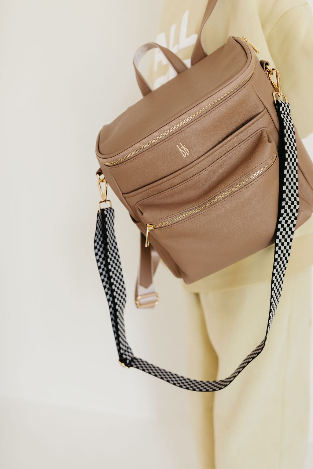 A person is holding the ff Diaper Bag in Taupe by forever french baby. This beige accessory, made from vegan leather, includes multiple zippered compartments and a checkered strap. It features gold hardware and a small monogram logo on the front pocket, with insulated bottle pockets for added convenience. The person complements the bag with a light-colored outfit.