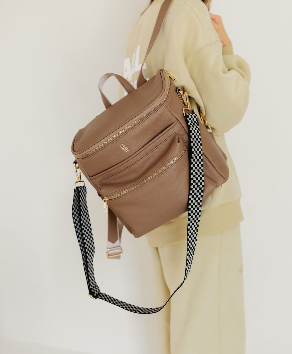 Dressed in beige attire, a person carries the stylish ff Diaper Bag in Taupe by Forever French Baby. This bag is crafted from vegan leather and features multiple pockets. It includes a black and white checkerboard shoulder strap, gold zippers, and insulated bottle pockets, perfectly blending functionality with fashion.