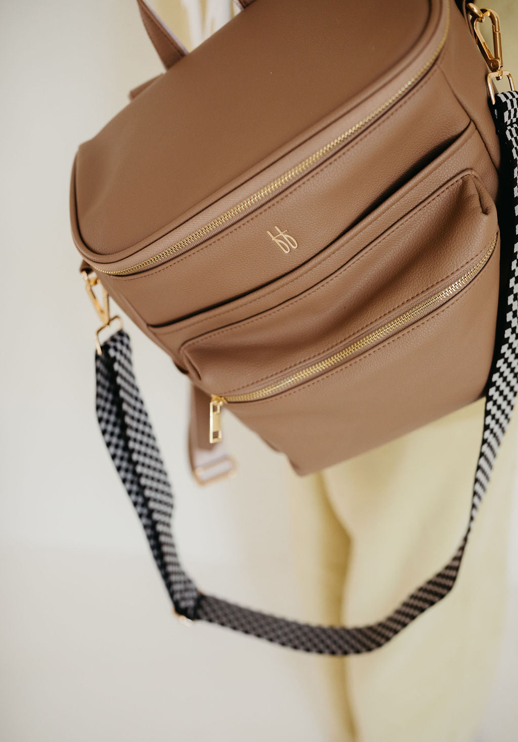 The ff Diaper Bag in Taupe from forever french baby hangs against a light background. This tan vegan leather backpack showcases gold zippers, multiple pockets including insulated bottle pockets, and a black and white checkered shoulder strap. The front pocket is embossed with a gold monogram.