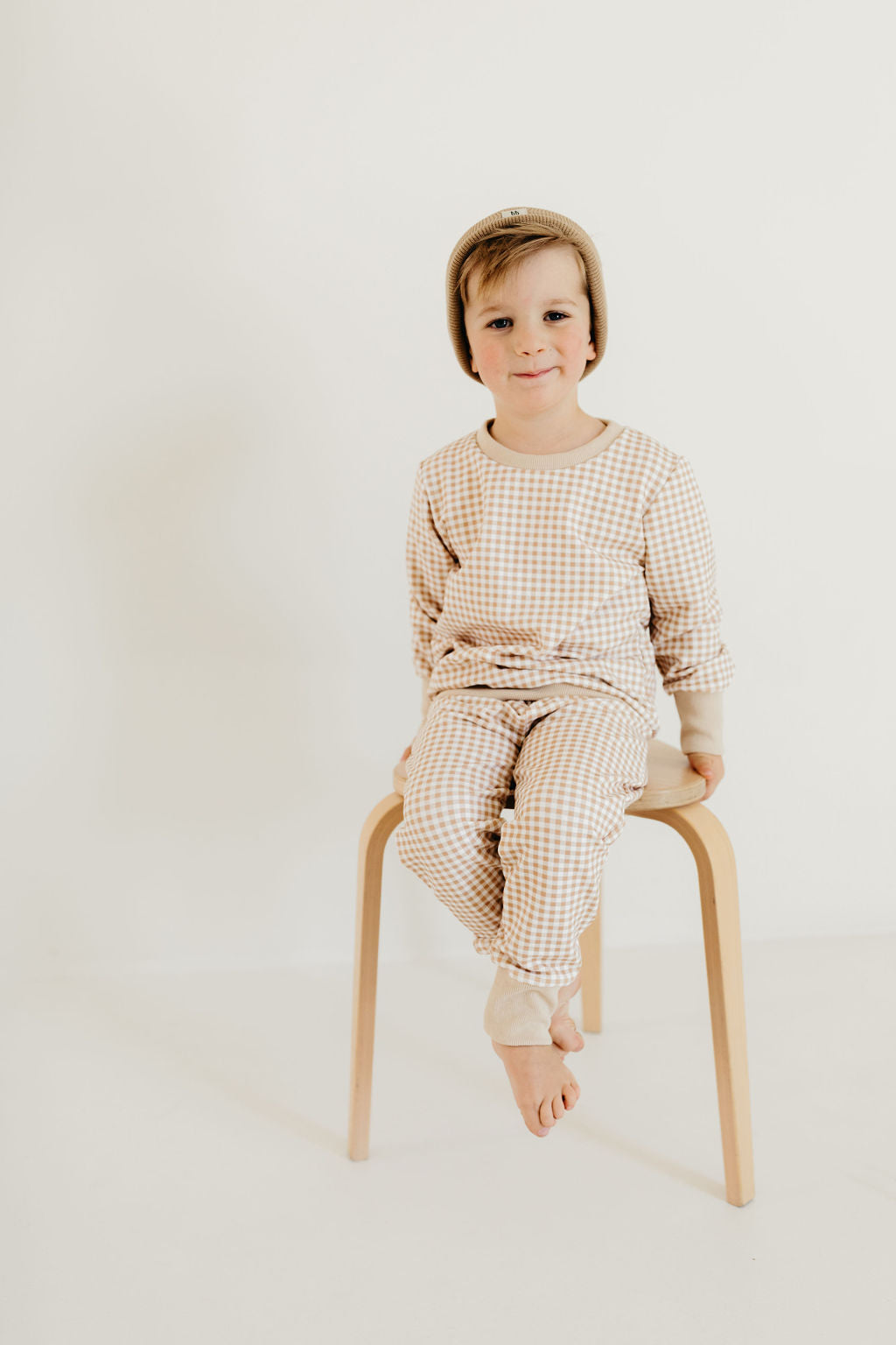 A young child perches on a wooden stool, dressed in the "Goldie Gingham" sweat set from forever french baby, featuring matching beige and white checkered pajamas with cuffed joggers and a cozy beanie. The plain, light background enhances the minimalist atmosphere. With a slight smile and bare feet, the scene exudes warmth and simplicity.