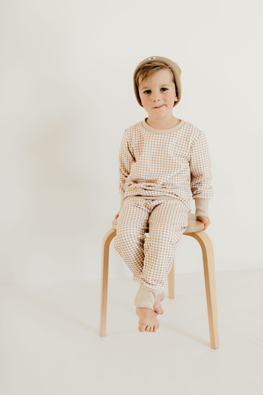 A young child dressed in the "Goldie Gingham" sweat set by forever french baby, featuring beige and white checkered cuffed joggers and a matching beanie, sits with feet crossed on a wooden stool against a light background.