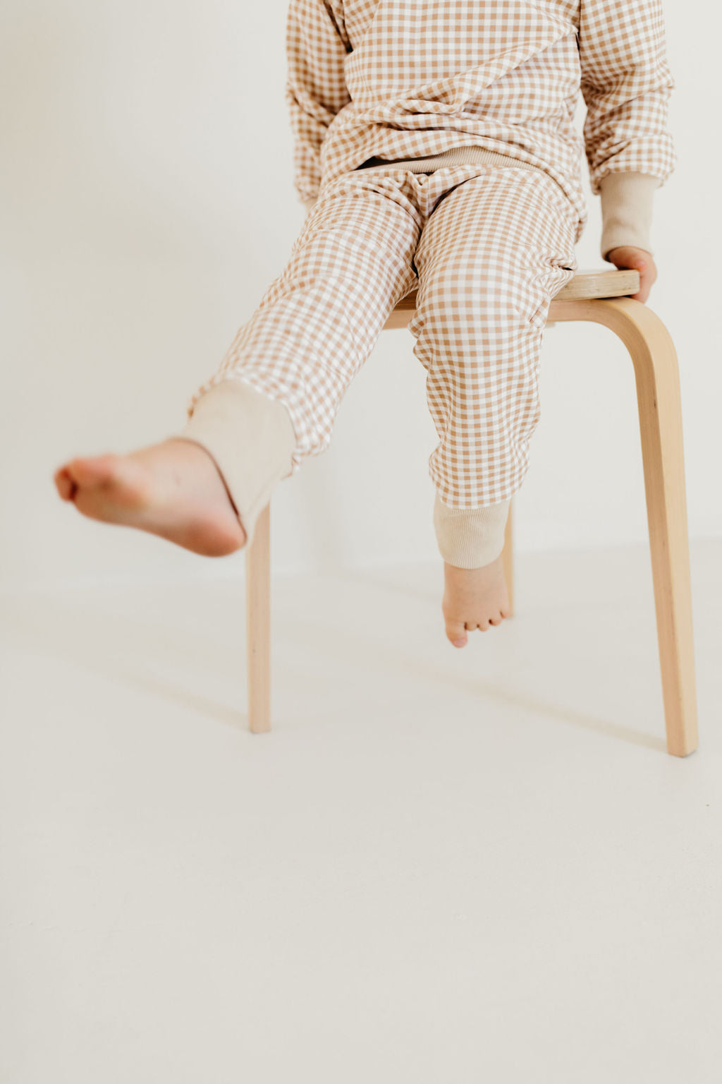 A child sits on a wooden stool, dressed in the Goldie Gingham Sweat Set by forever french baby. One leg stretches out while the other dangles off the edge, set against a soft, neutral background.