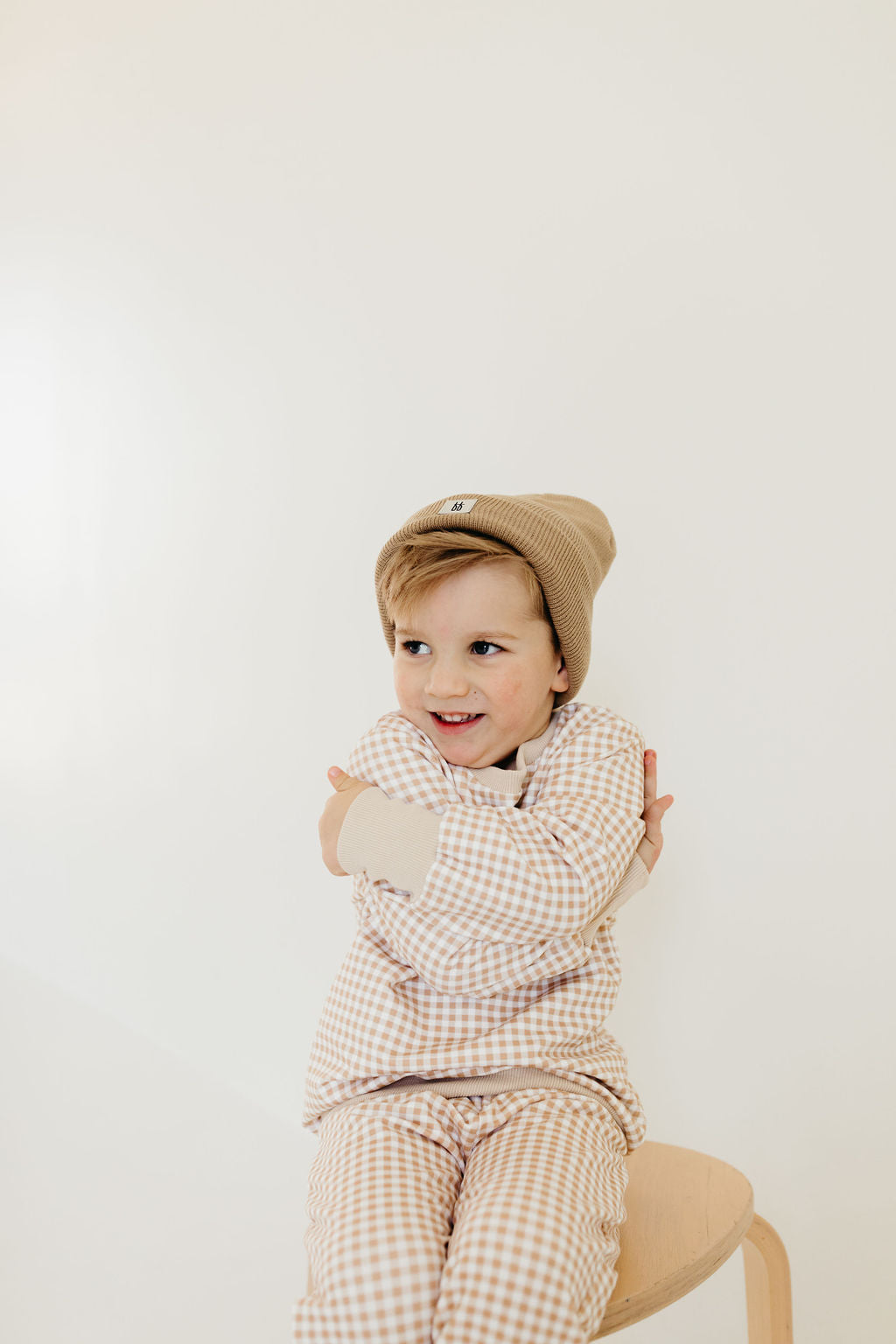 A young child, dressed in the Sweat Set | Goldie Gingham from forever french baby and a cozy crewneck sweater, sits on a wooden stool beneath a tan knit beanie. They are smiling and hugging themselves, exuding warmth against the plain, light-colored wall.