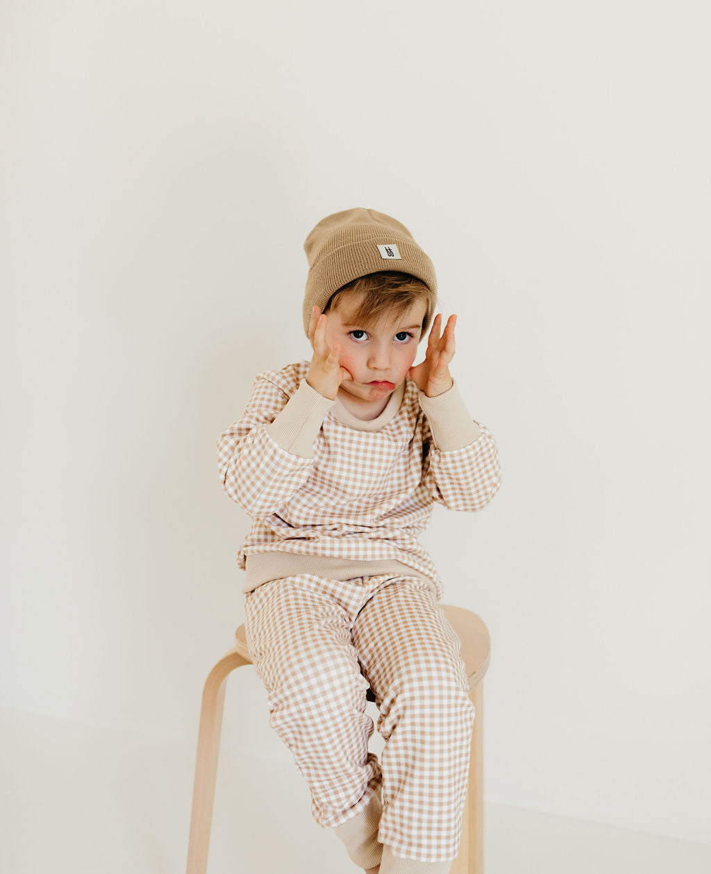 A young child in a cozy crewneck sweater and Goldie Gingham outfit by forever french baby, complete with a matching beanie, sits on a wooden stool against a plain background, playfully holding their hands to their face.