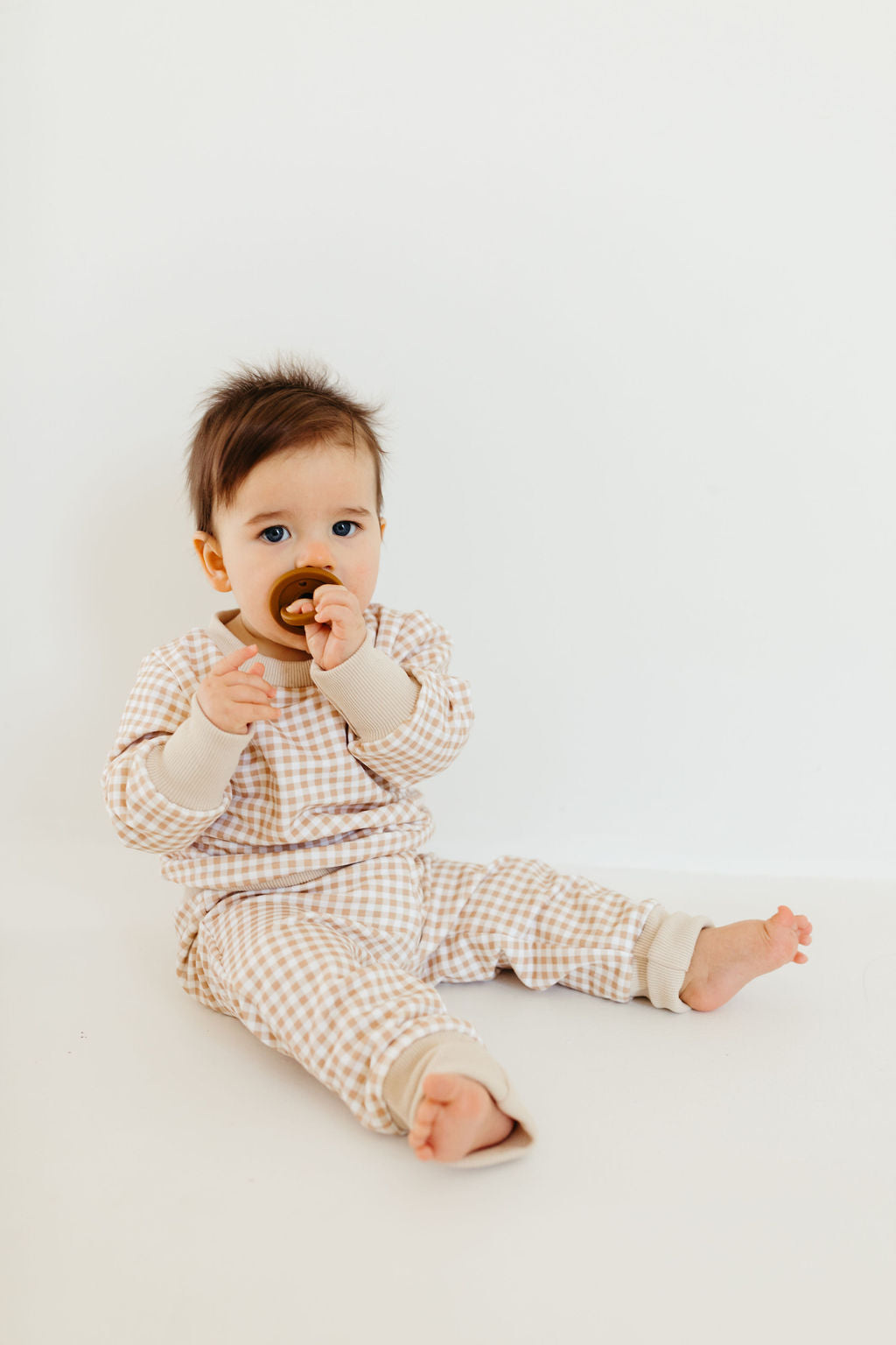 A baby sits on a white background wearing the Sweat Set | Goldie Gingham from forever french baby. The outfit features a beige and white checkered design, paired with cuffed joggers. The baby holds a brown pacifier in its mouth, with bare feet and a curious expression.