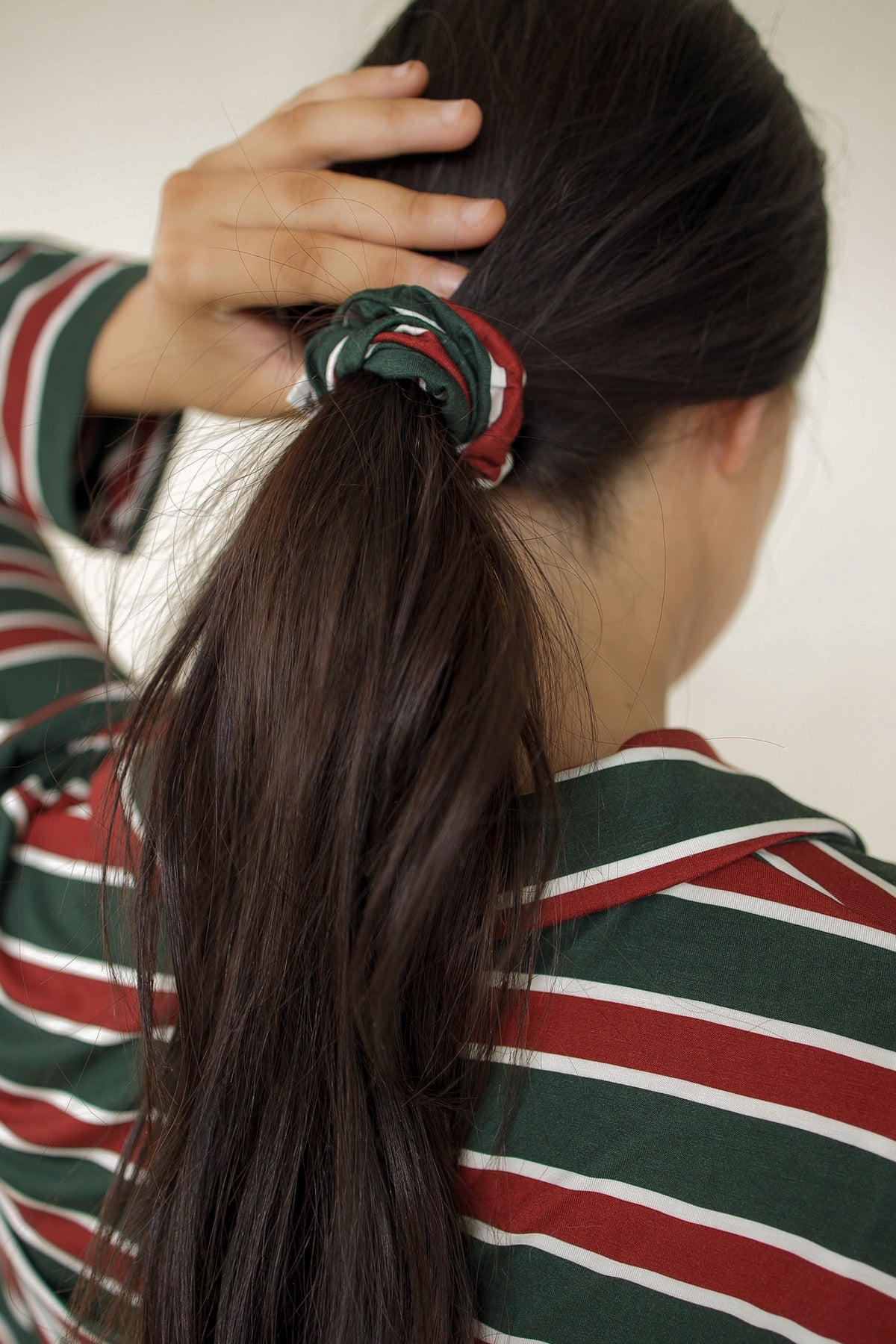 A person with long dark hair worn in a ponytail secured by a forever french baby Scrunchie | Holiday Stripe.