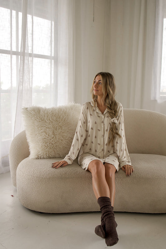 A woman wearing a Women’s Bamboo Sleeping Dress | Gingersnap sits on a sofa, dressed in the long-sleeved, button-down bamboo nightgown with a gingerbread man print and dark brown socks.