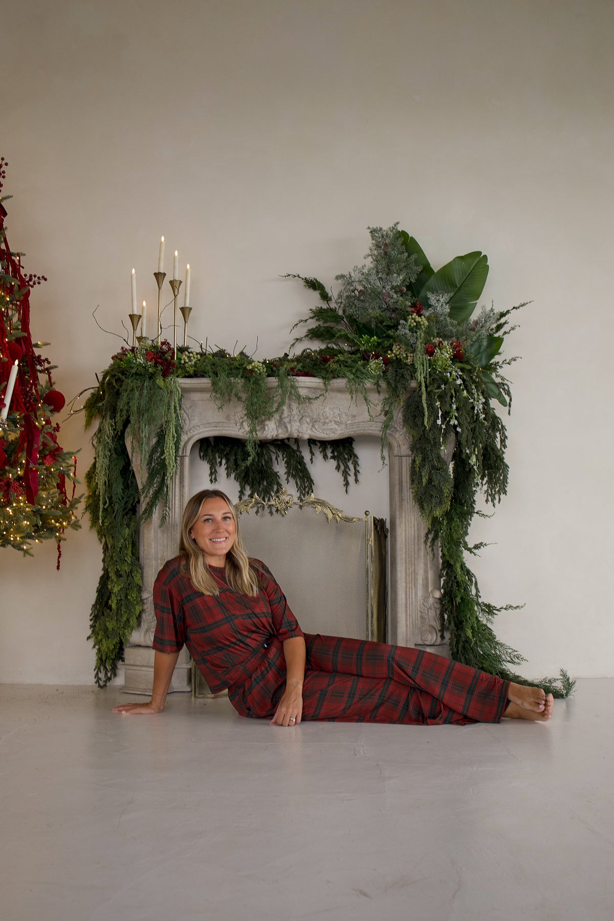 A woman wearing a Women’s Boxy Tee Lounge Set | Christmas Plaid in red and black sits on the floor in front of a fireplace decorated with greenery and candles.