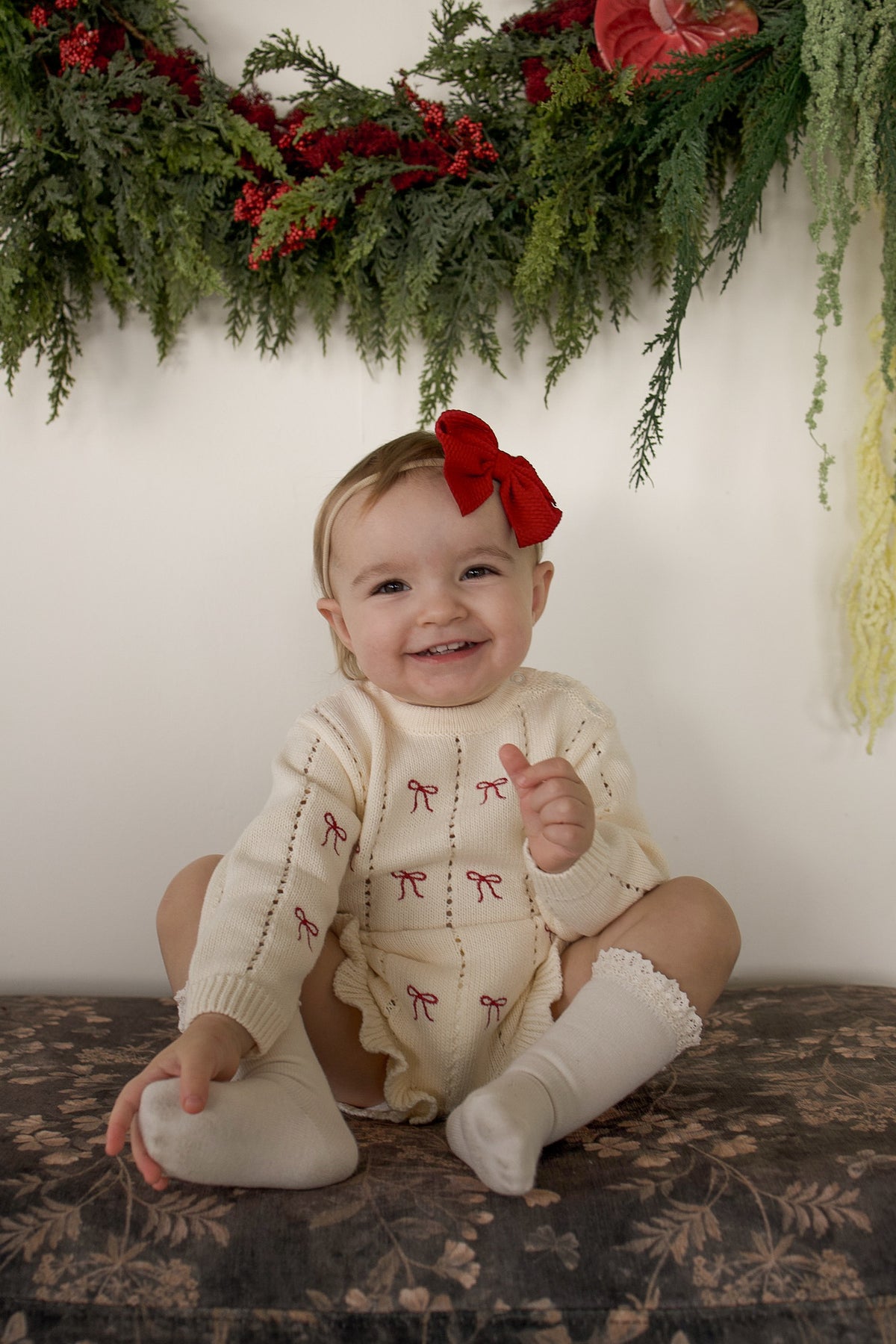 A smiling baby wearing a Knit Ruffle Romper | Candy Cane, styled with a matching red hair bow and lace-trimmed socks, sitting in front of a holiday greenery garland.