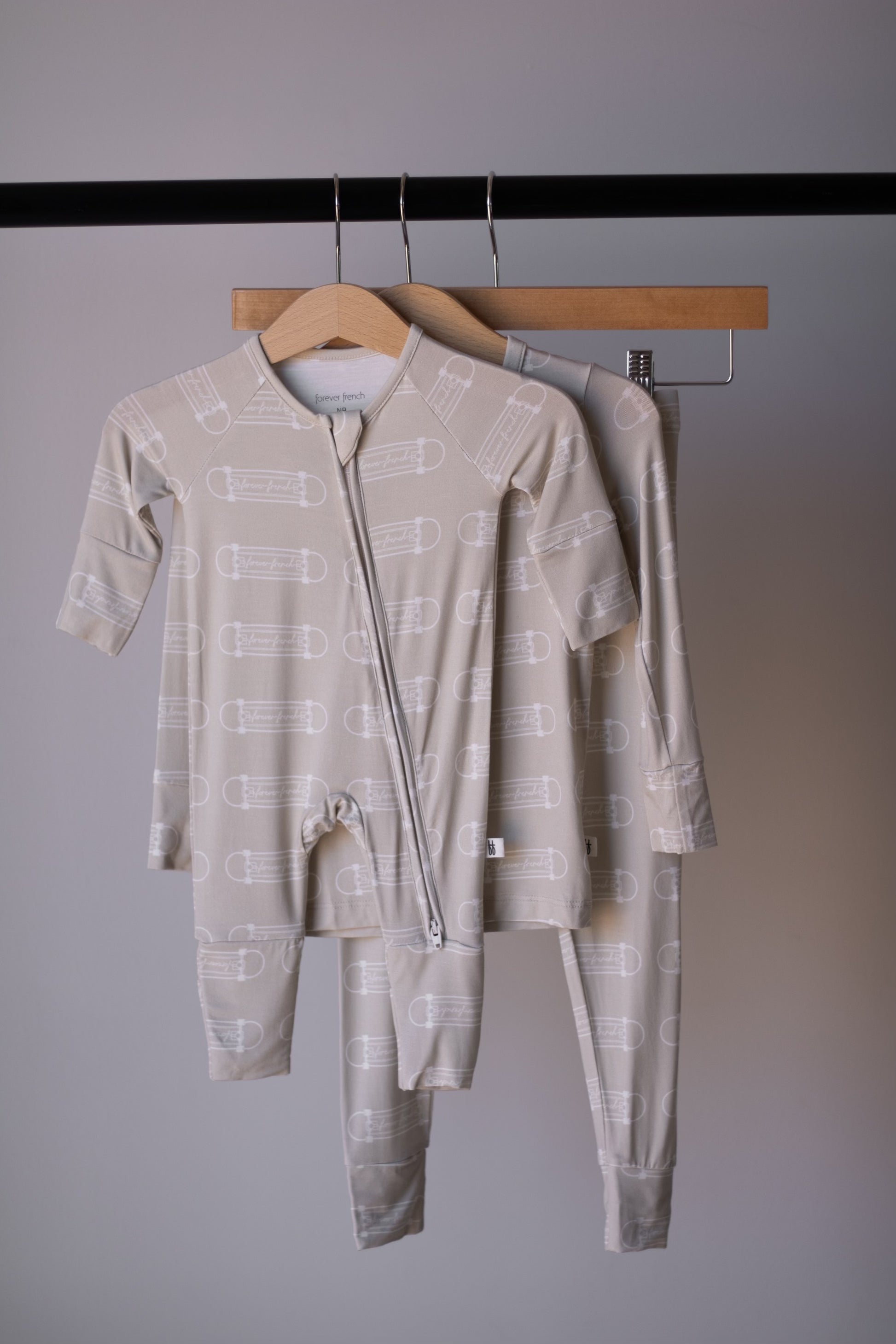Two sets of forever french baby Bamboo Zip Pajamas | Sk8er Boy in light beige with white safety pin pattern hang on wooden hangers from a black rack against a gray wall, offering soft, breathable kids' sleepwear for restful nights.
