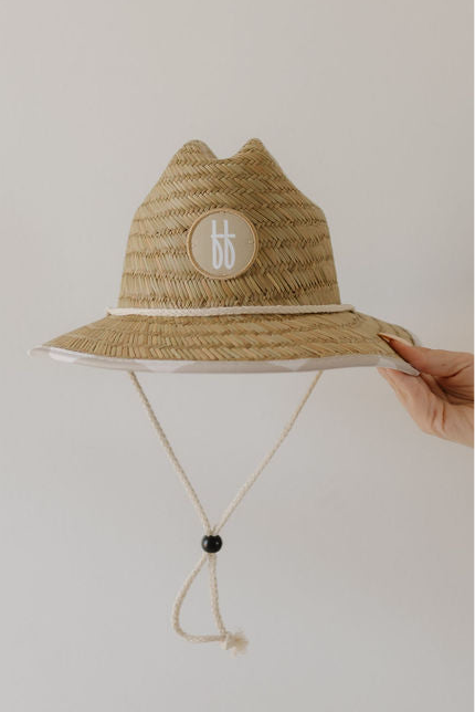 A hand holds the Straw Hat | Checkerboard by forever french baby, featuring a chin cord and a round "bb" patch on the front. Perfect for babies and children, set against a plain, light background.