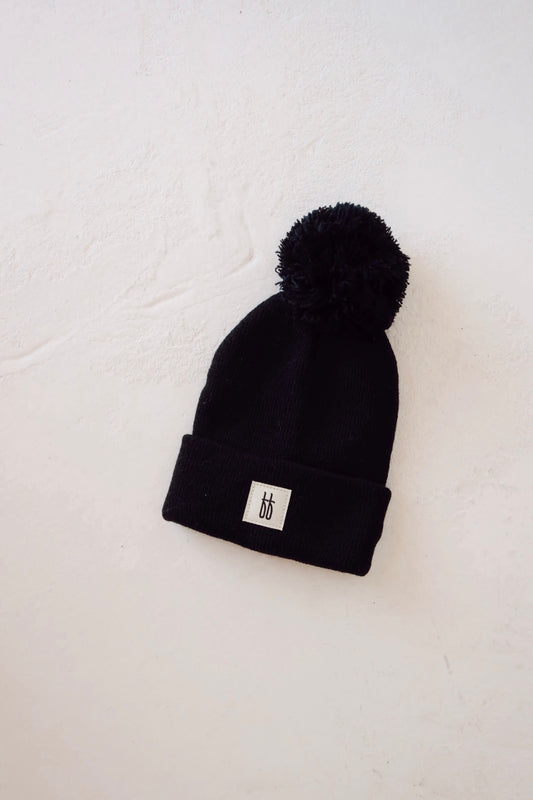 The ff Knit Pom Top Beanie in black by forever french baby is laid out on a light-colored surface. This cozy and stylish hat features a large pom-pom on top and a white rectangular patch with a small, minimalist symbol in black on its folded brim.