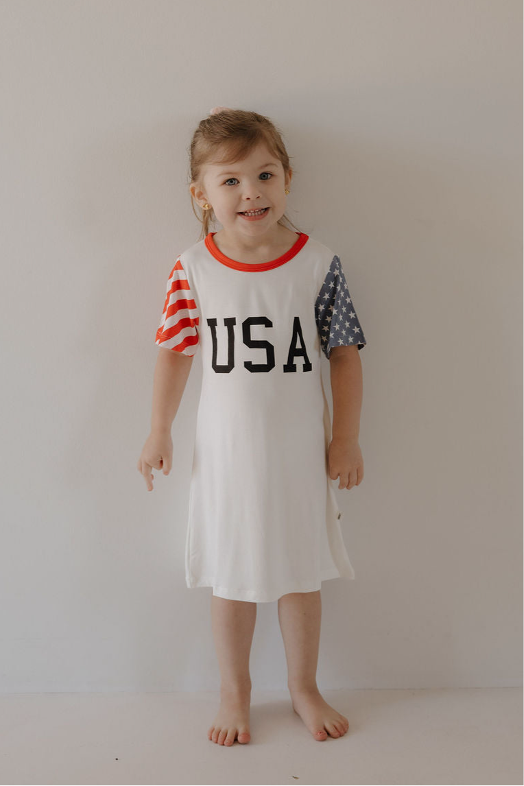 A young child wears the forever french baby Bamboo T-Shirt Dress | Stars & Stripes, featuring "USA" on the front, one red striped sleeve, and one blue sleeve with white stars for a patriotic look.