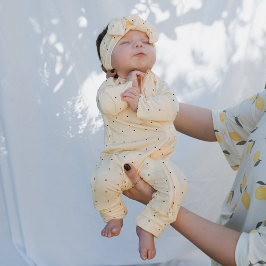 A baby in forever french baby's soft yellow Bamboo Zip Pajamas | Veda with a matching headband is held by an adult. Sunlight and leaf shadows move across the white sheet as the peaceful baby rests, eyes closed and hands together.