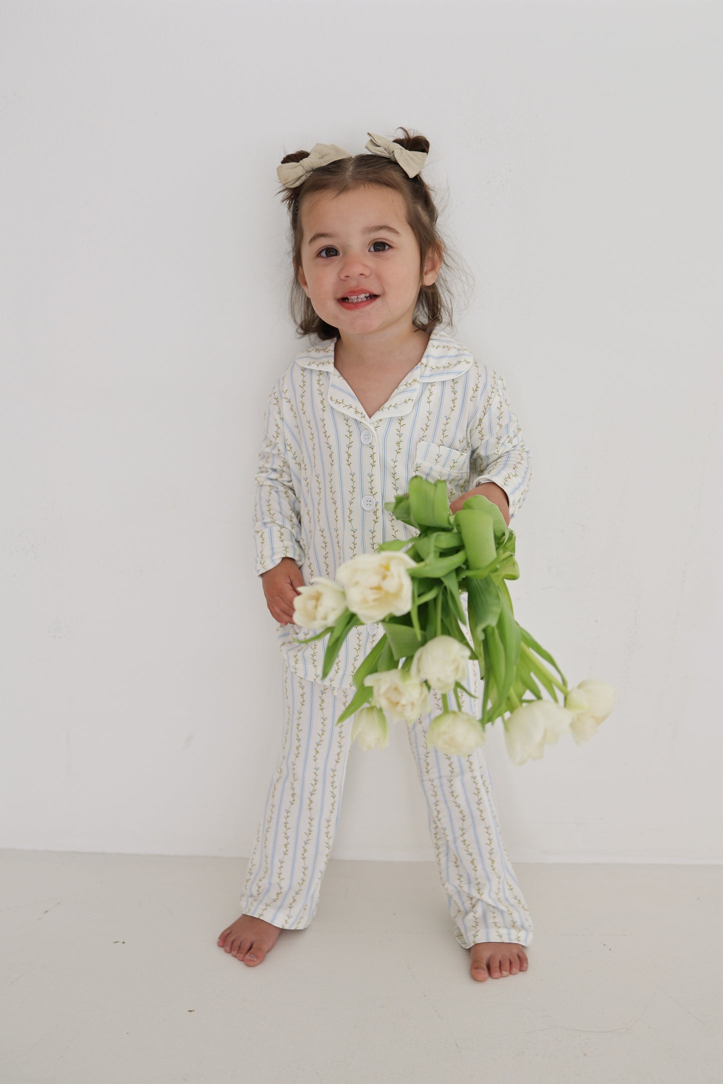 A young girl with brown pigtails and white bows smiles barefoot against a white wall, holding white tulips. She wears the Girl’s Bamboo Button Up Lounge Set in Dainty Vines by forever french baby.
