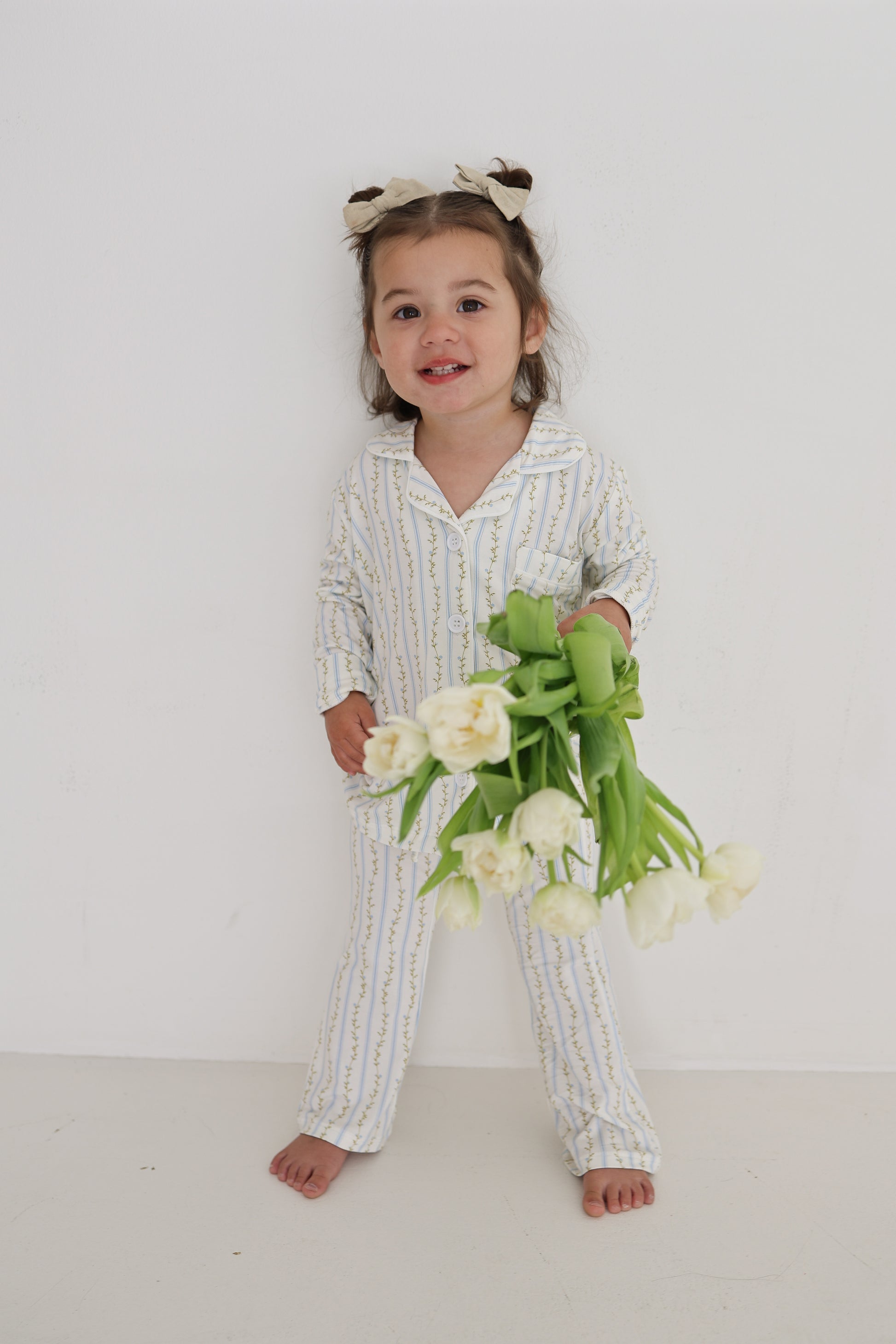 A young girl with brown pigtails and white bows smiles barefoot against a white wall, holding white tulips. She wears the Girl’s Bamboo Button Up Lounge Set in Dainty Vines by forever french baby.