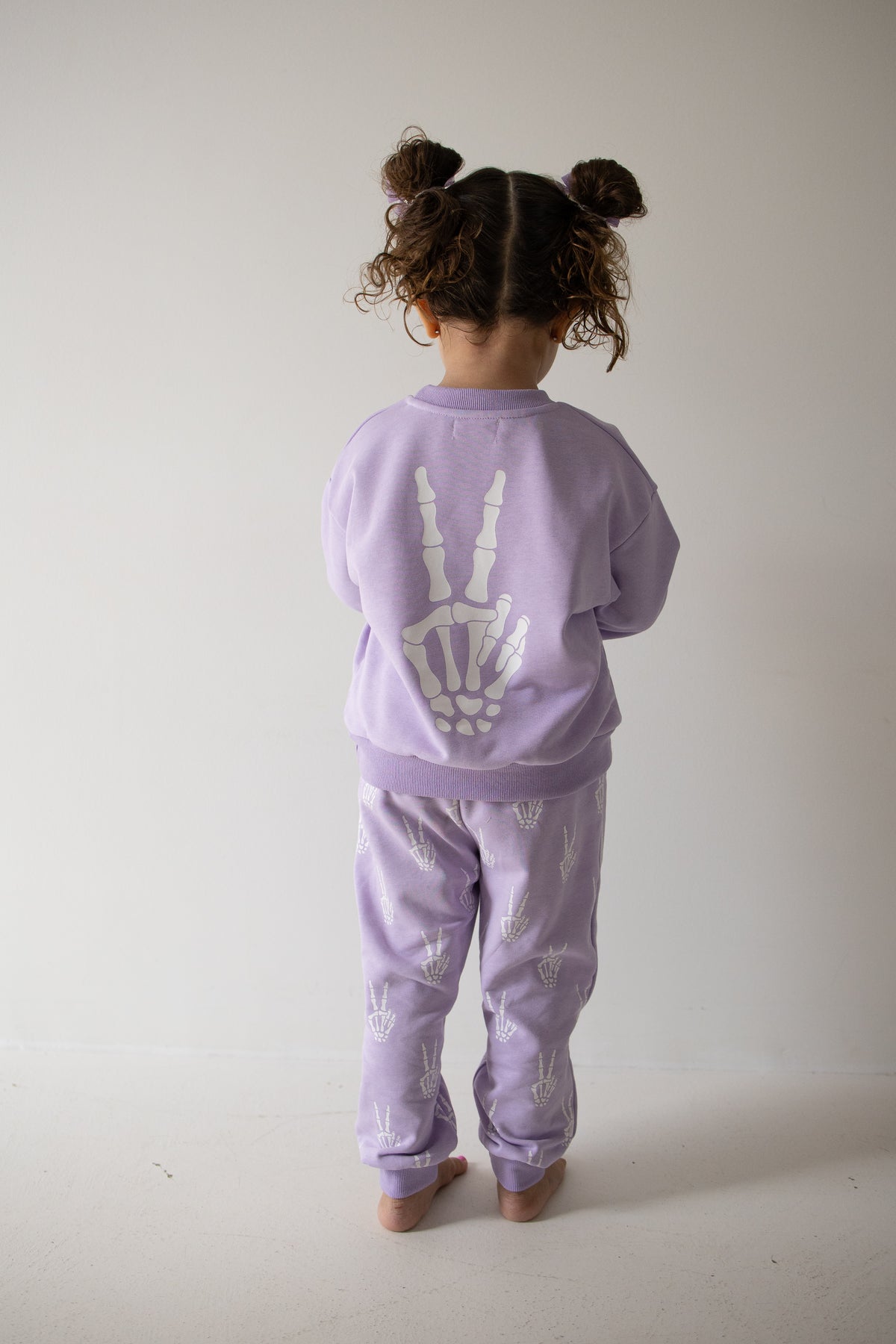 A young child with curly hair in pigtails stands barefoot, facing away, dressed in the forever french baby Child Sweat Set | Enchanted Skelly—a light purple set featuring white skeletal hand designs on the back and legs—perfect for everyday adventures.