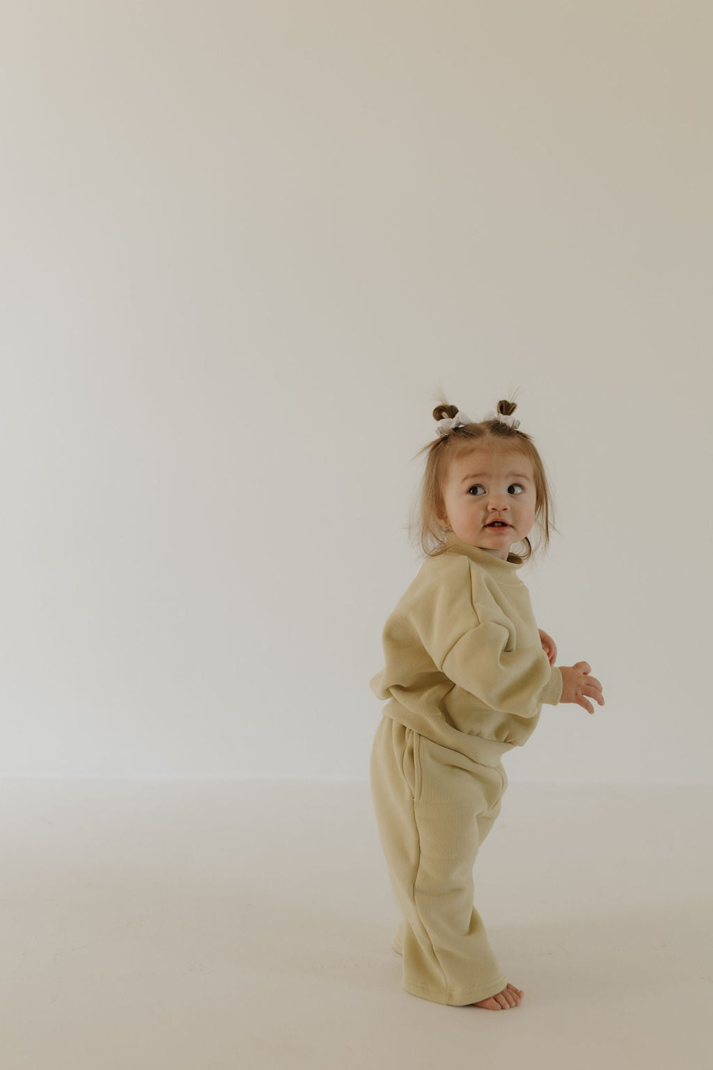 A young child stands barefoot on a light background, wearing the matching tan "Child Sweat Set | All the Good Things" from forever french baby. The child has small pigtails and looks curiously over their shoulder. The setting is simple and minimalistic.