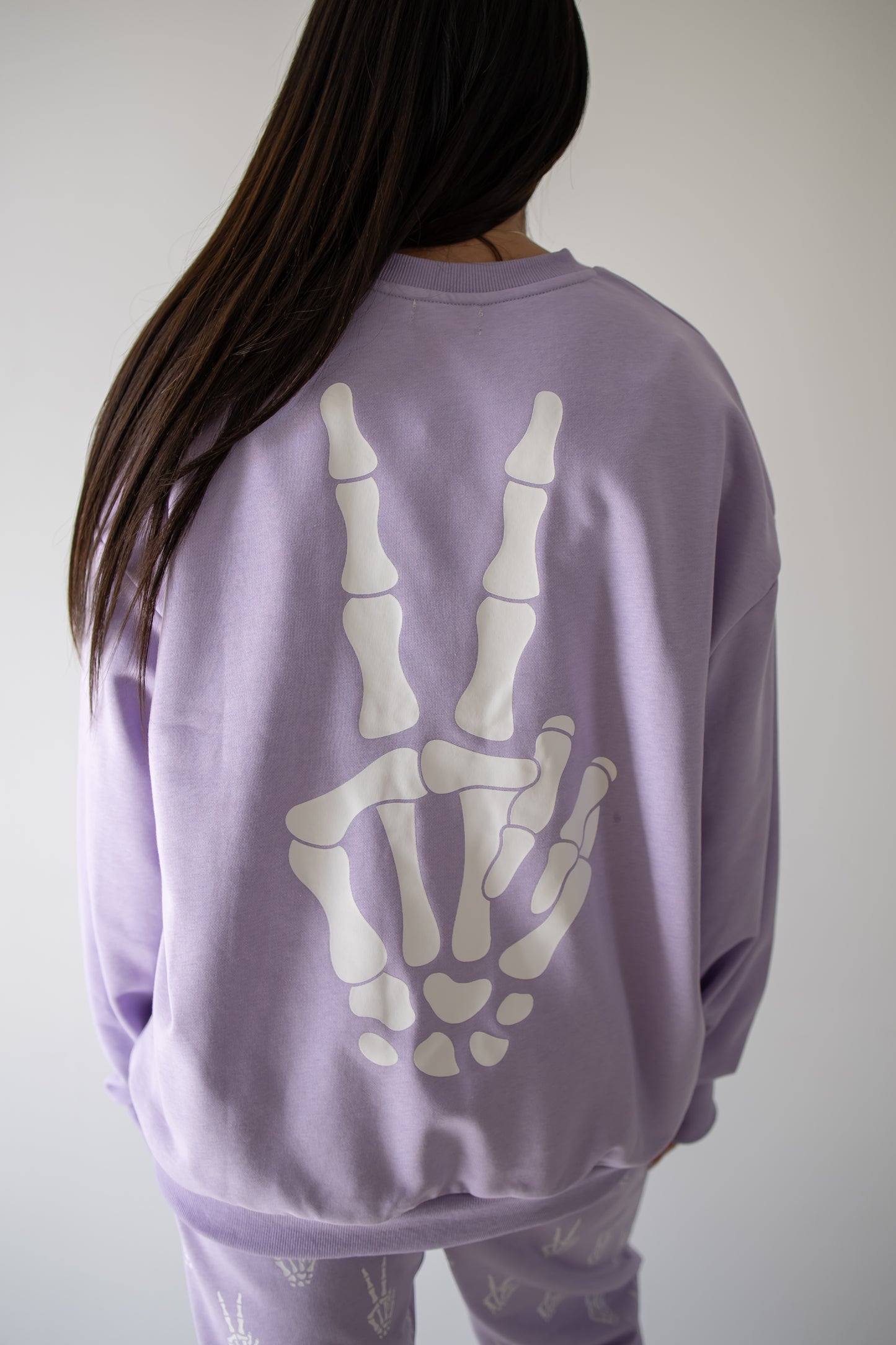 A woman with long dark hair models the forever french baby Women’s Sweat Set | Enchanted Skelly in lavender, featuring a bold white skeletal hand peace sign graphic on the back and coordinating pants adorned with small white accents—an ideal spooky season ensemble—standing against a simple backdrop.